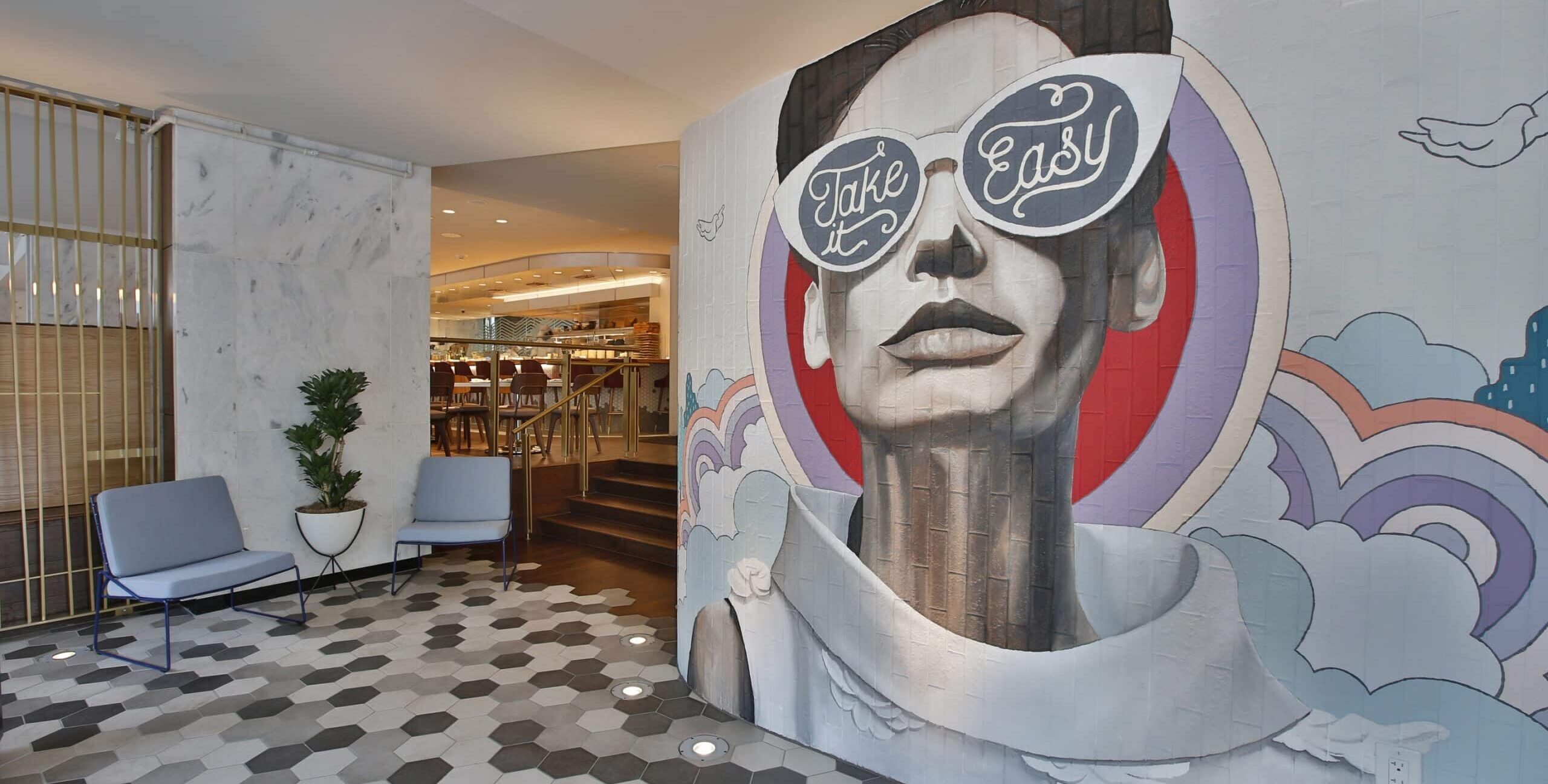 A modern interior with a large mural of a person wearing oversized sunglasses that read “Take it Easy.” The wall features colorful abstract shapes behind the portrait. The floor has hexagonal tiles in gray, white, and black. Two minimalist chairs and a potted plant sit against a marble wall with vertical gold bars, and a warmly lit dining area is visible in the background.