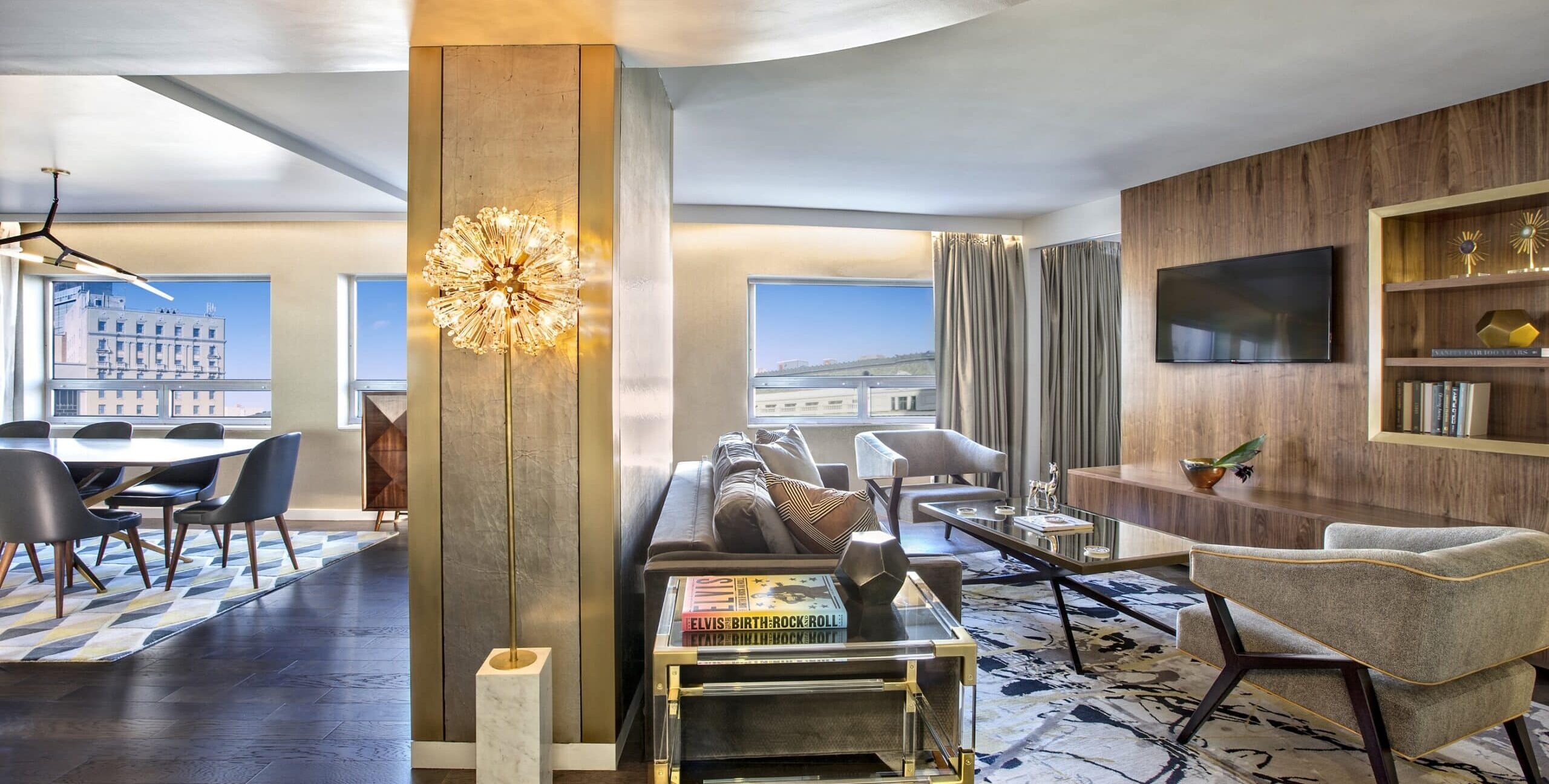 A contemporary living and dining space with a gray sectional sofa, two gray armchairs, and a wooden coffee table on a patterned rug. A wood‑paneled wall with built‑in shelves and a mounted TV sits behind the seating area. The dining area features a rectangular table with dark gray chairs on a geometric rug. A tall gold decorative floor lamp stands between the two spaces, and large windows show buildings outside.