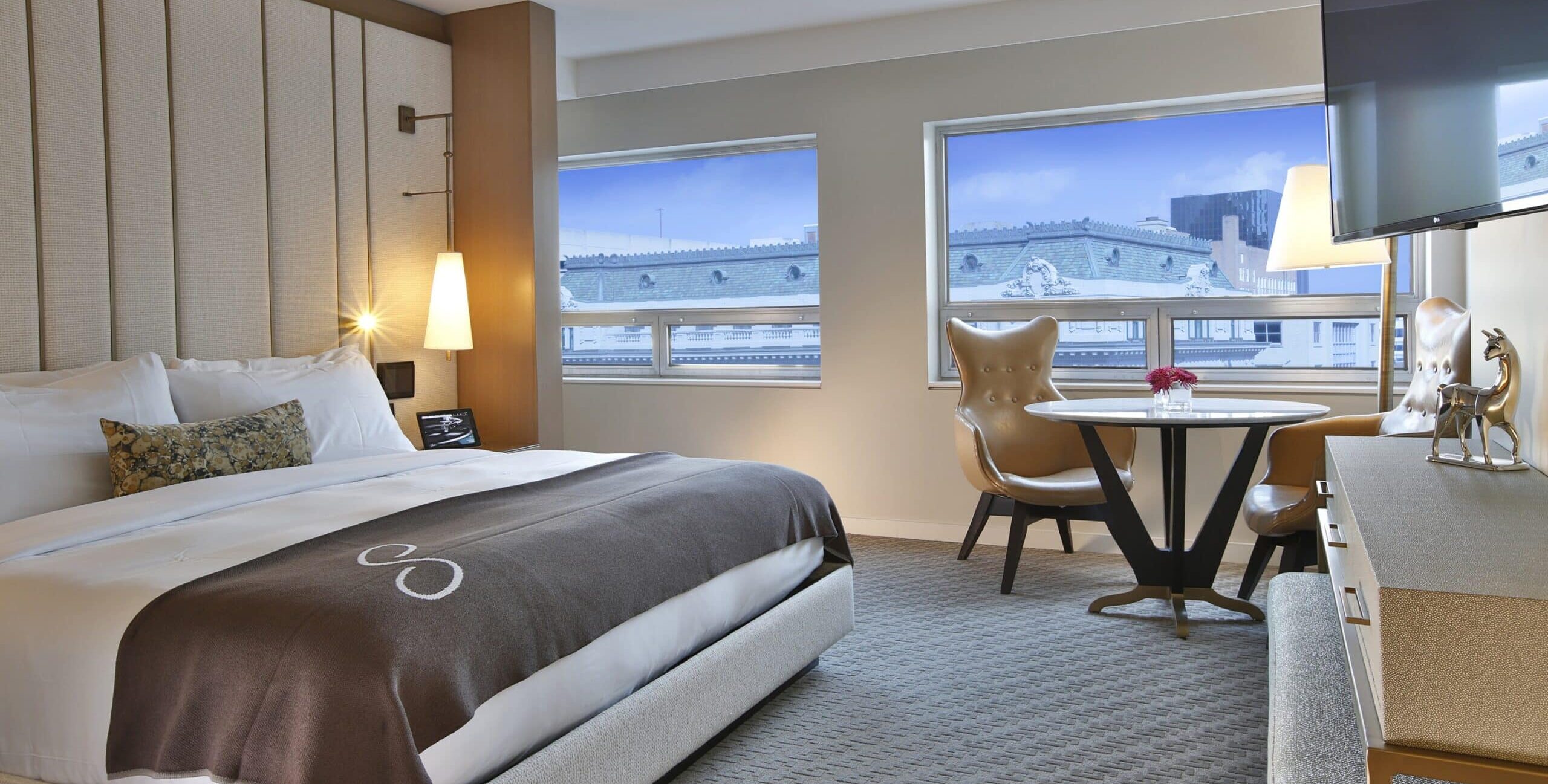 A modern hotel room with a large bed dressed in white linens, a patterned brown blanket, and an accent pillow. Two pendant lights hang beside the upholstered headboard. A round table with two tan leather chairs sits in front of large windows overlooking ornate rooftops. A flat‑screen TV is mounted above a light‑colored dresser with decorative items.