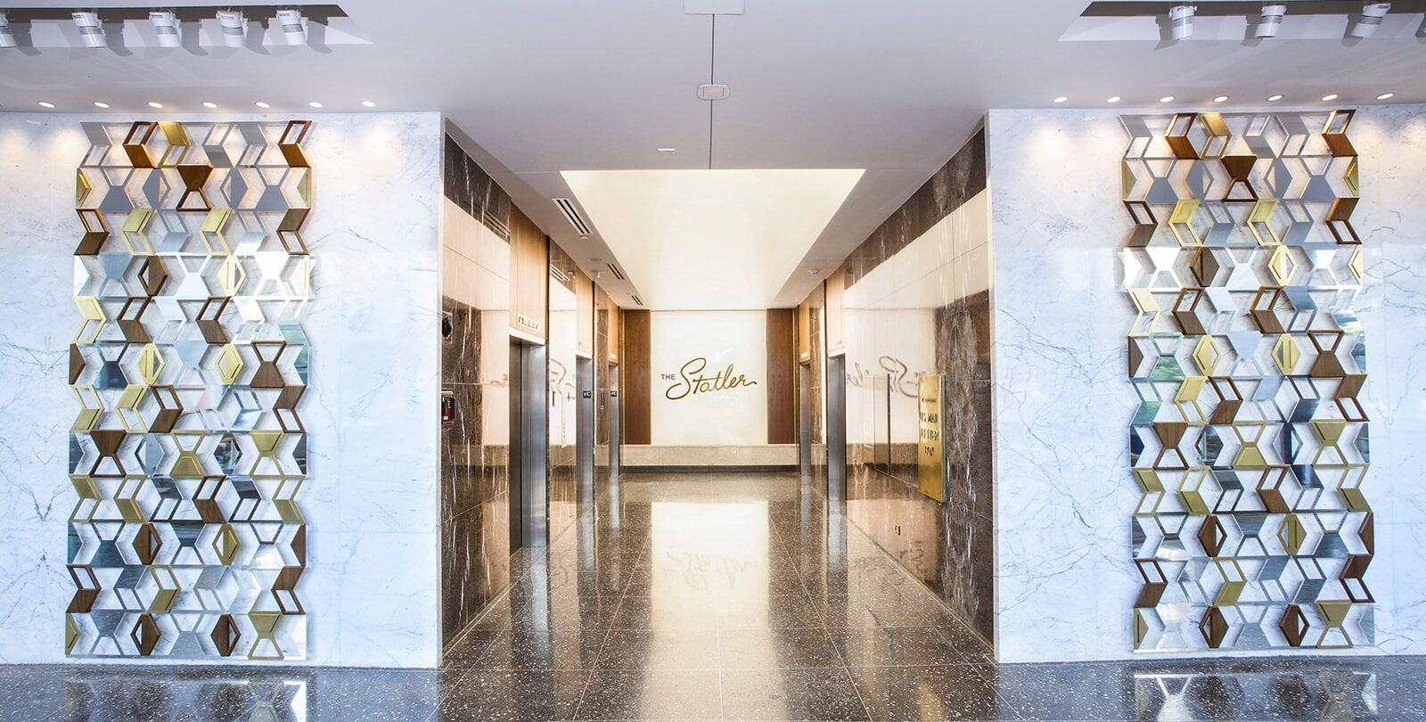 A sleek elevator lobby with polished dark terrazzo floors, white marble walls, and hexagonal metallic decorative panels. Two elevator doors are framed in wood and dark stone. At the end of the hallway, a wall sign reads “The Statler” in cursive script.