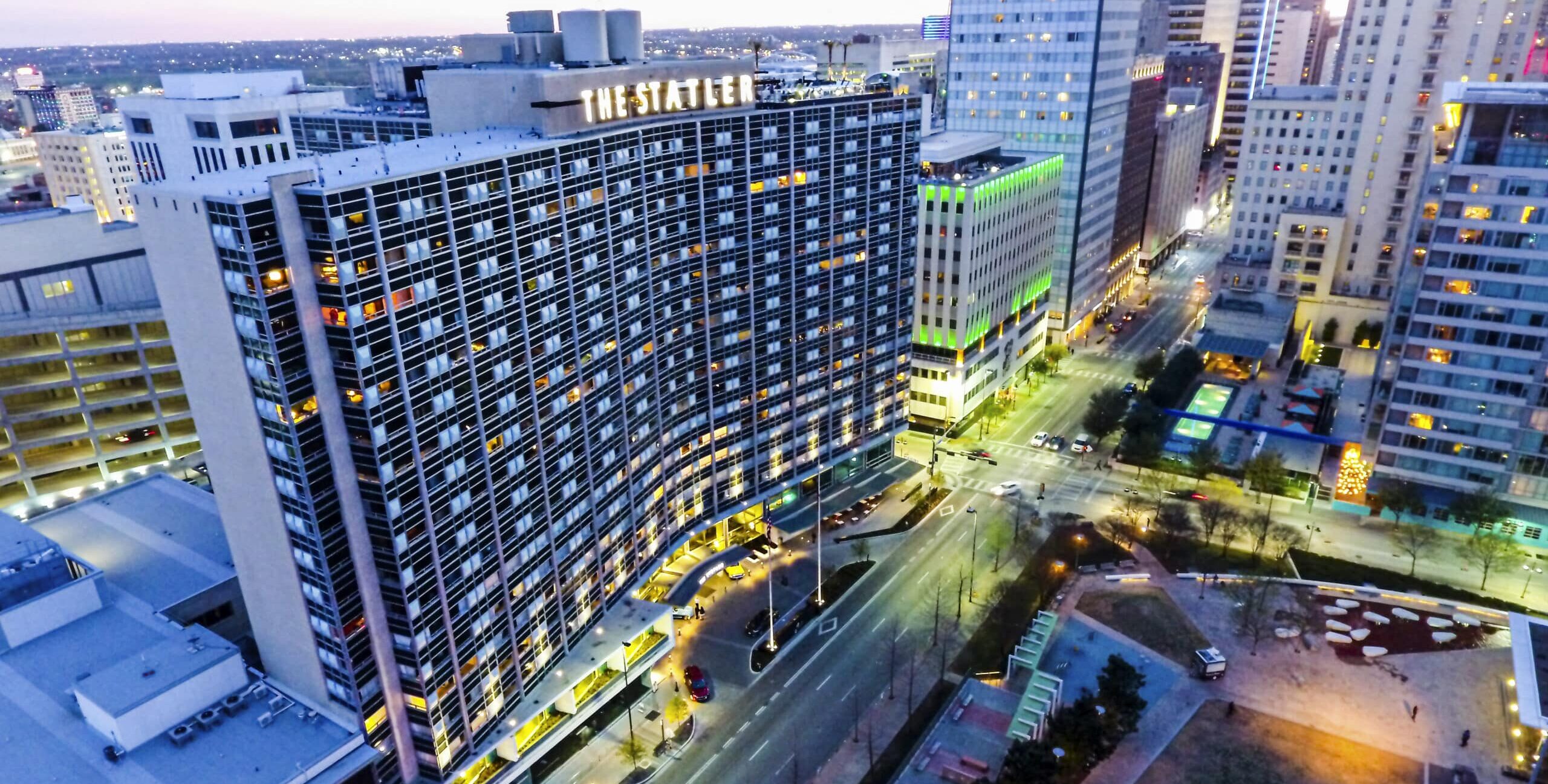An aerial view of a downtown area at dusk, centered on a curved building with illuminated rooftop letters reading “THE STATLER.” Surrounding high‑rises display colorful lighting, and streets below are busy with cars and pedestrians. A small plaza with pathways and a lit Christmas tree appears in the lower right.