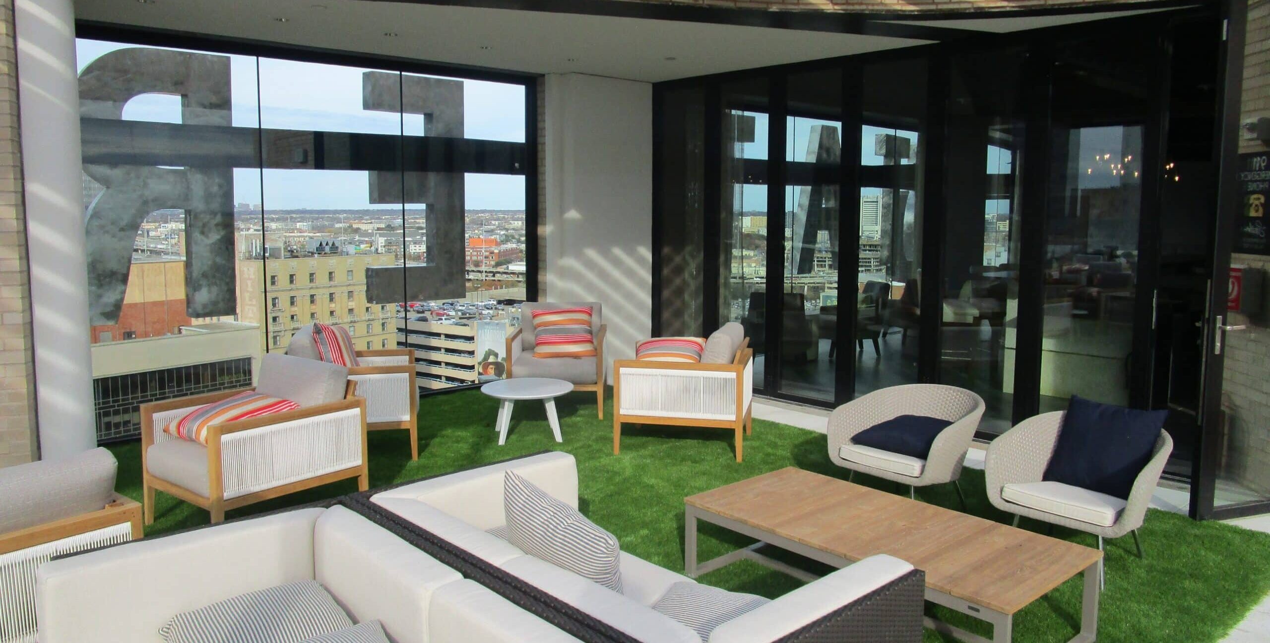 A rooftop terrace with cushioned wicker chairs, sofas, and wooden coffee tables arranged on artificial grass. A wooden pergola provides partial shade. Large glass doors open to an indoor space, and a city skyline is visible in the distance.