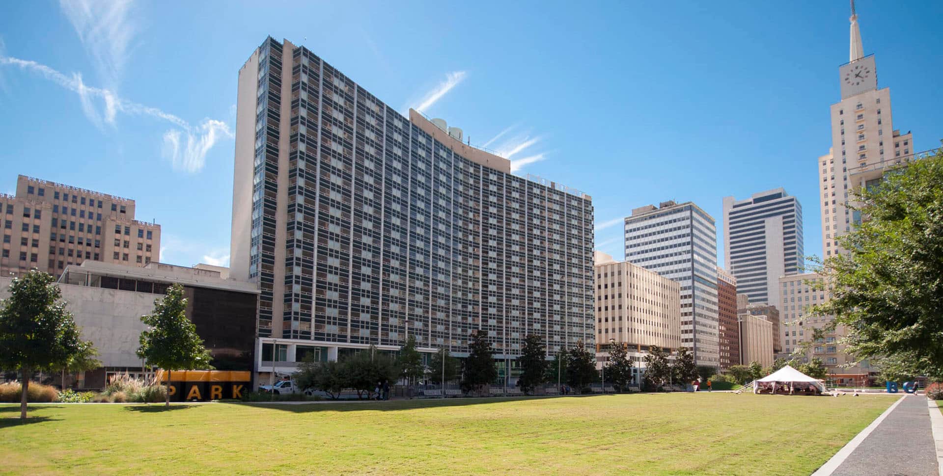A grassy park area bordered by trees with several tall office buildings and a clock tower in the background under a clear blue sky.