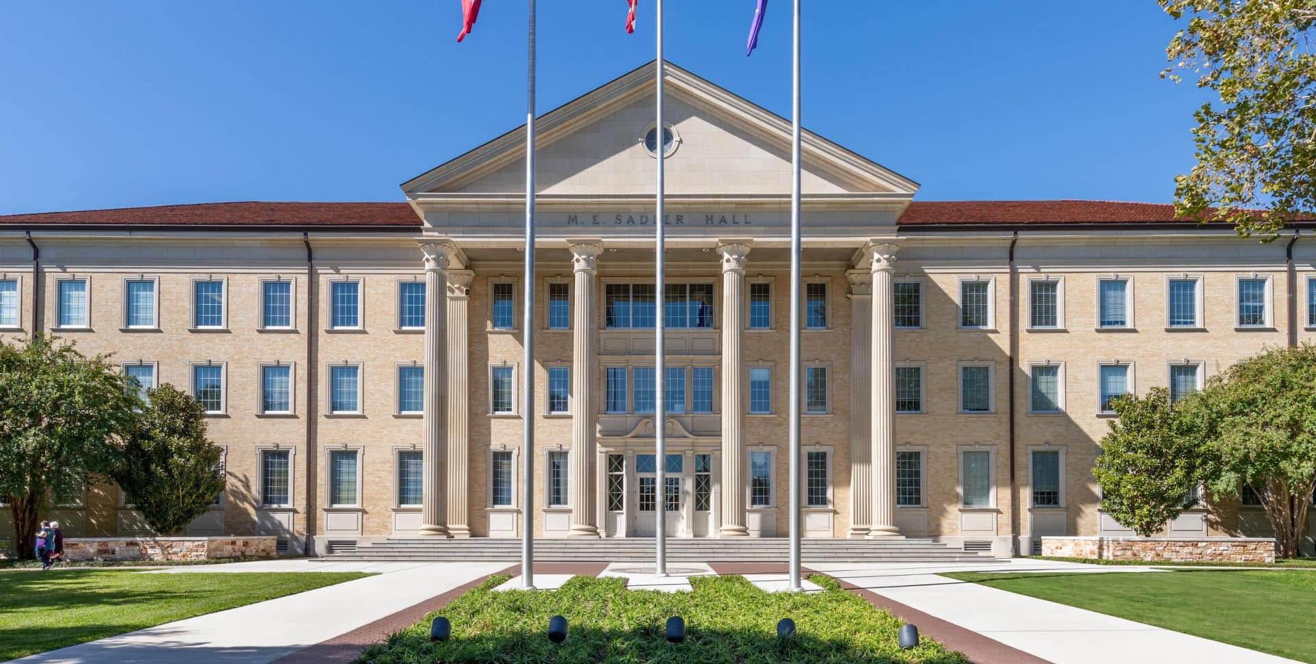 A large, tan brick academic building with tall columns, three flagpoles in front, and manicured lawns under a clear blue sky.