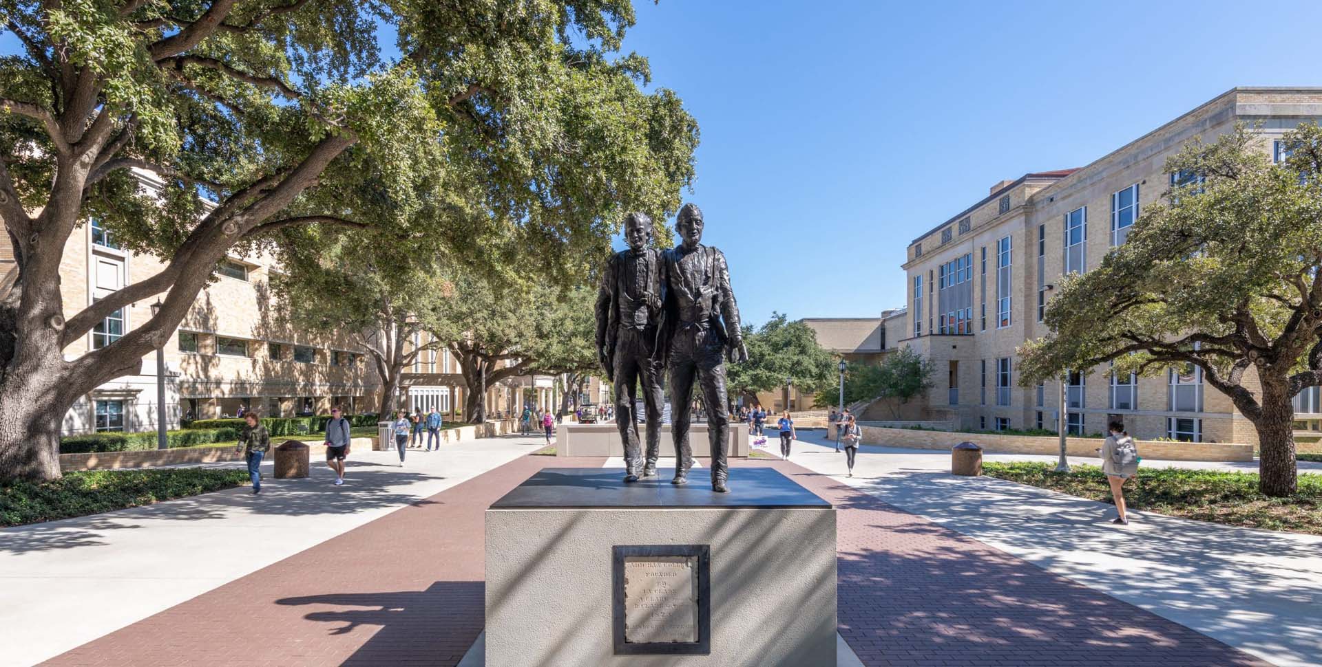 A bronze statue of two men stands on a pedestal in a paved walkway, surrounded by trees and campus buildings with people walking nearby.