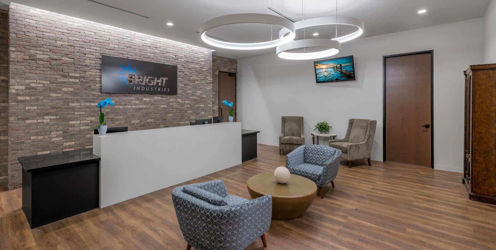 A modern office reception area with a front desk, two blue armchairs, two gray chairs, a round coffee table, and a wall sign reading "Bright Industries.