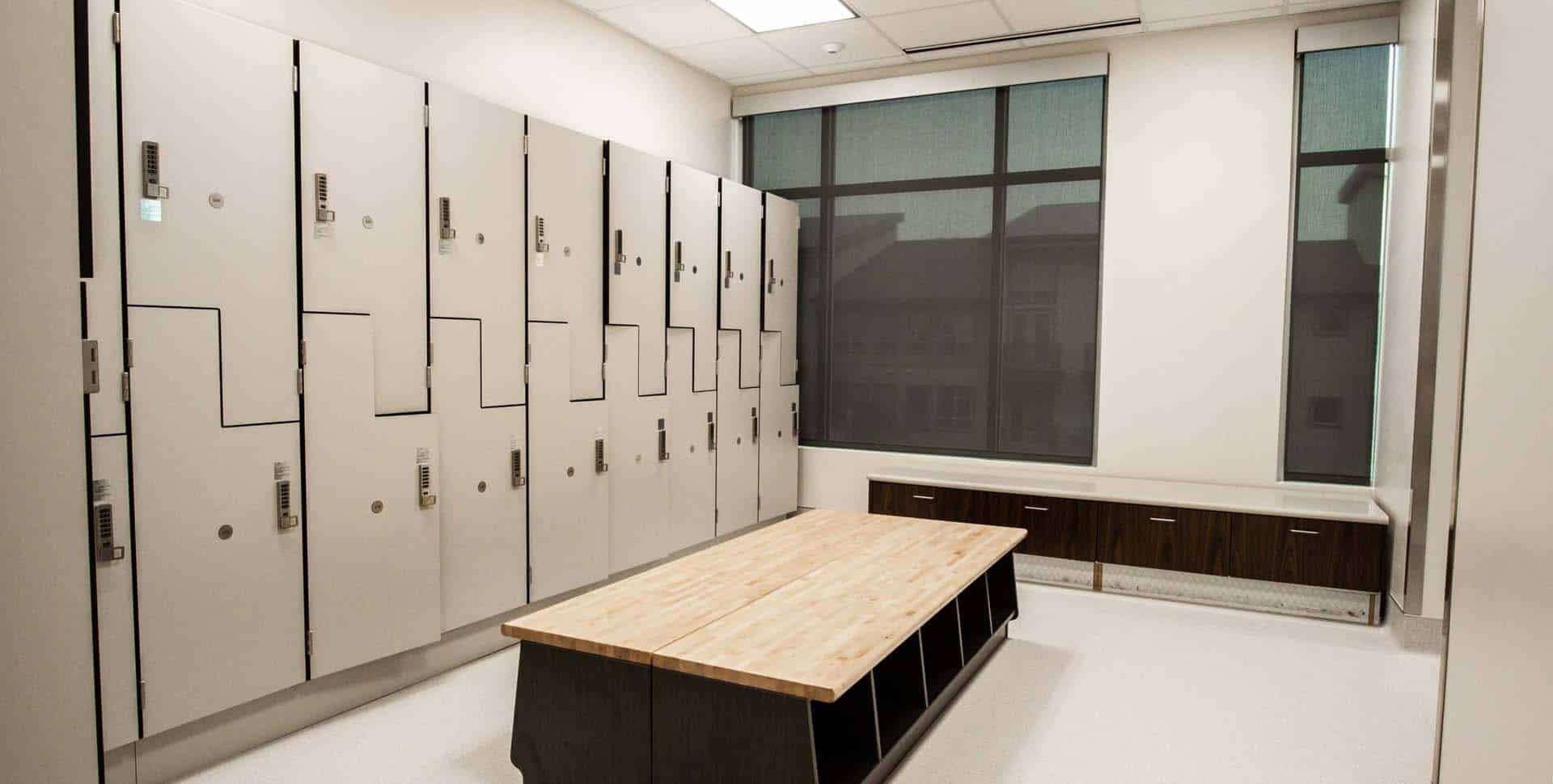A modern locker room with white lockers, a wooden bench in the center, and large windows letting in natural light.