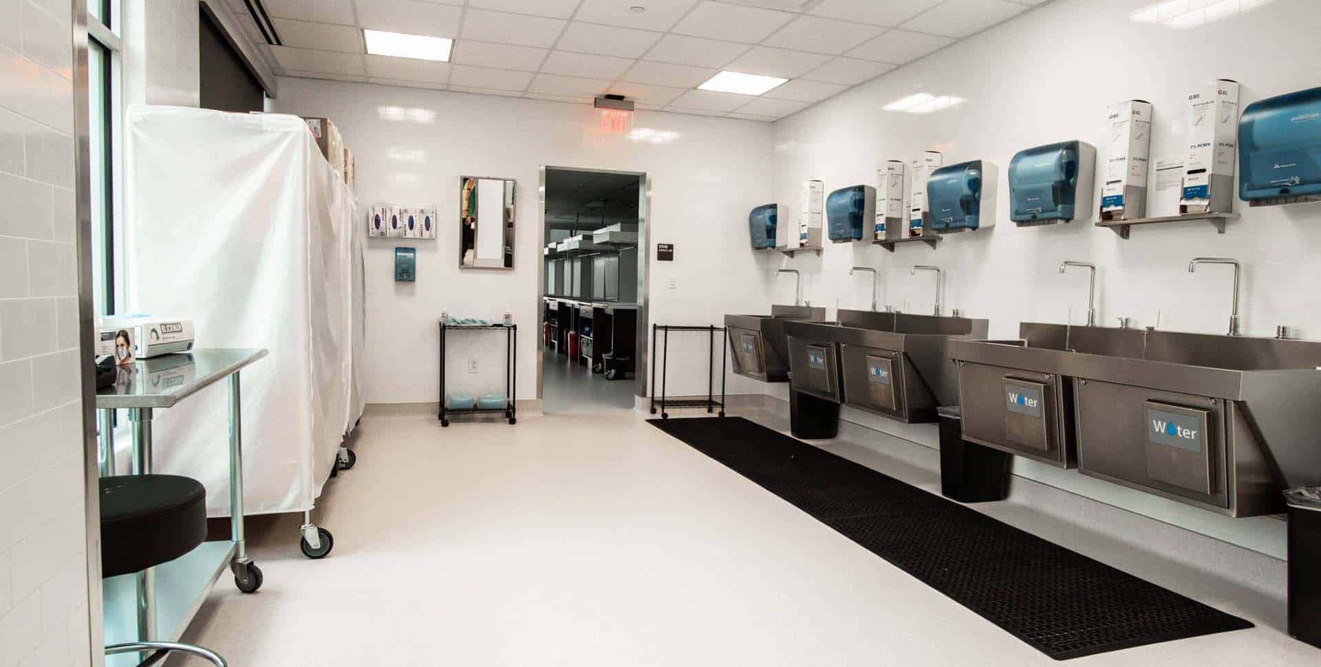 A clean, well-lit room with multiple stainless steel sinks, wall-mounted soap and paper towel dispensers, and a covered cart, likely in a medical or laboratory setting.