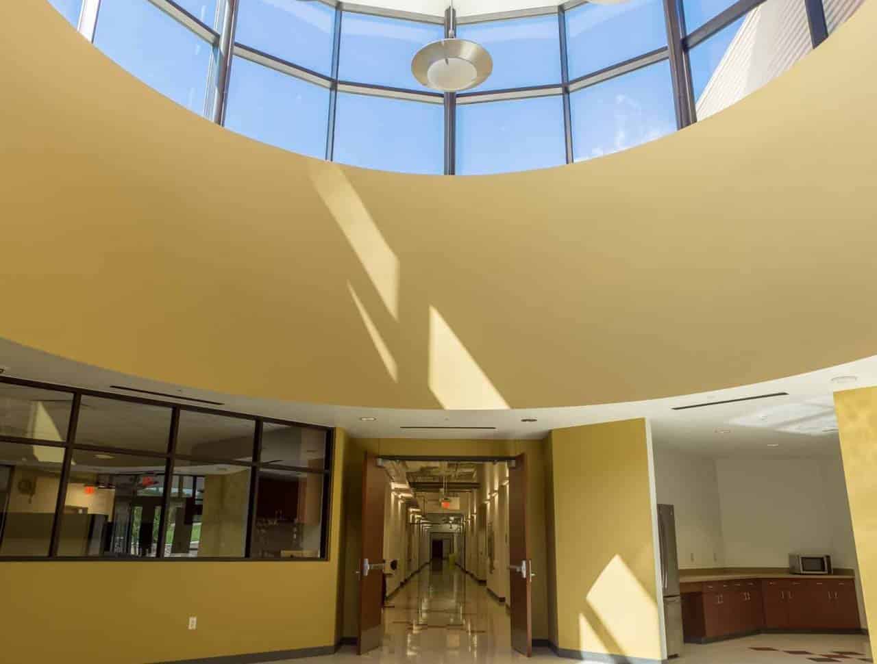 Sunlit hallway with a circular skylight, yellow walls, polished floor, and glass windows on the left. Doors line the corridor; cabinets and appliances are on the right.