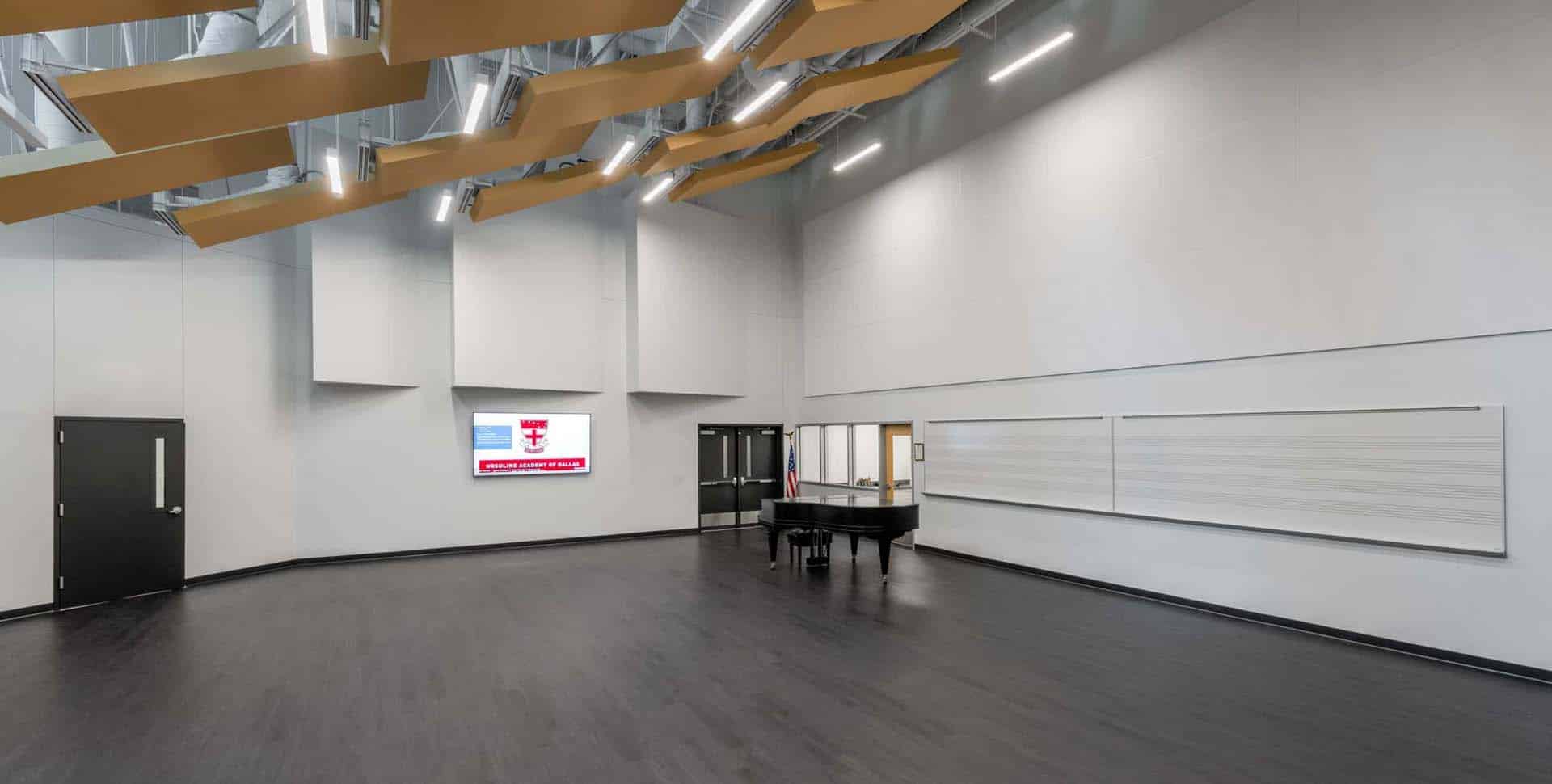 A modern music rehearsal room with a black piano, wall-mounted whiteboards with staff lines, a digital display, and acoustic panels on the ceiling.