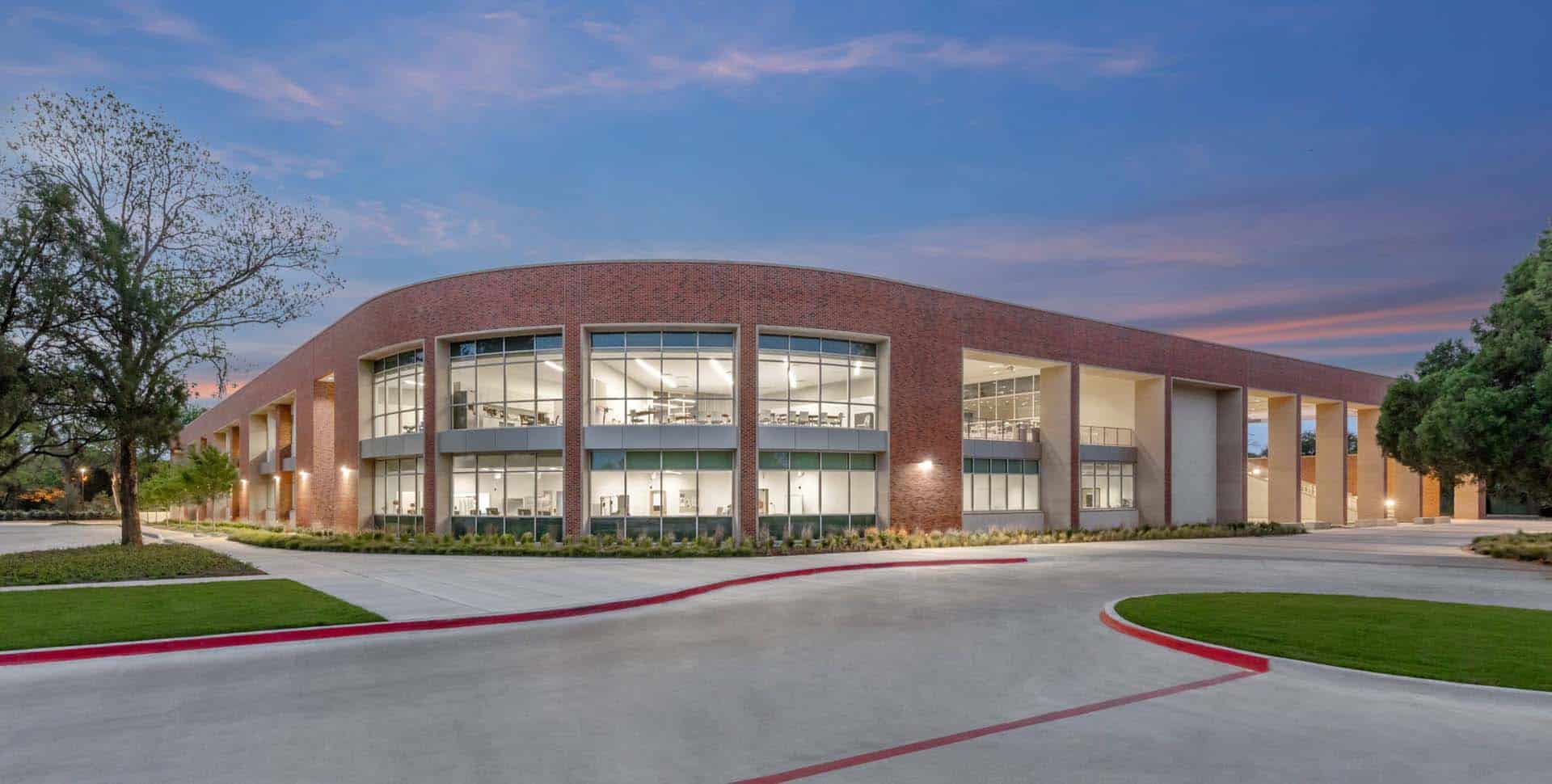 Modern two-story brick building with large windows and curved design, surrounded by landscaped greenery and a paved driveway at dusk.