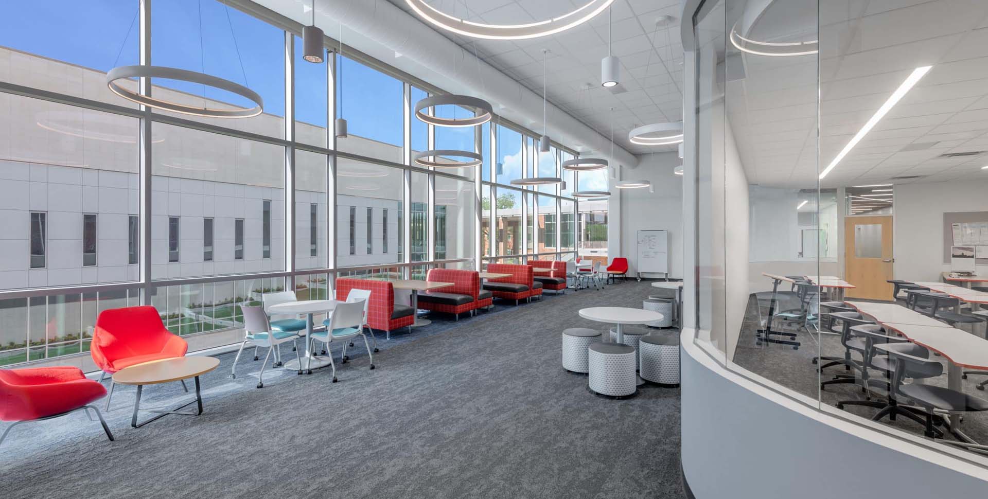 Modern office lounge with large windows, red and white seating, round tables, and circular ceiling lights next to a glass-walled meeting room.