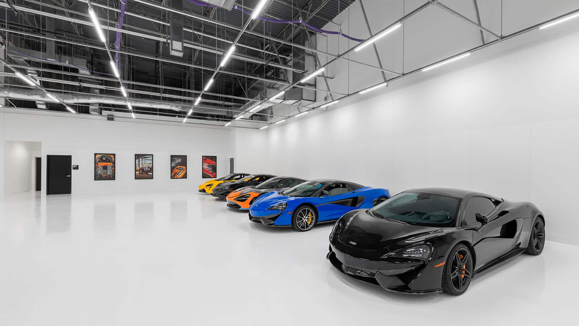 A row of colorful sports cars, including black, blue, orange, yellow, and gray models, parked in a bright, modern showroom with white walls and ceiling lights.