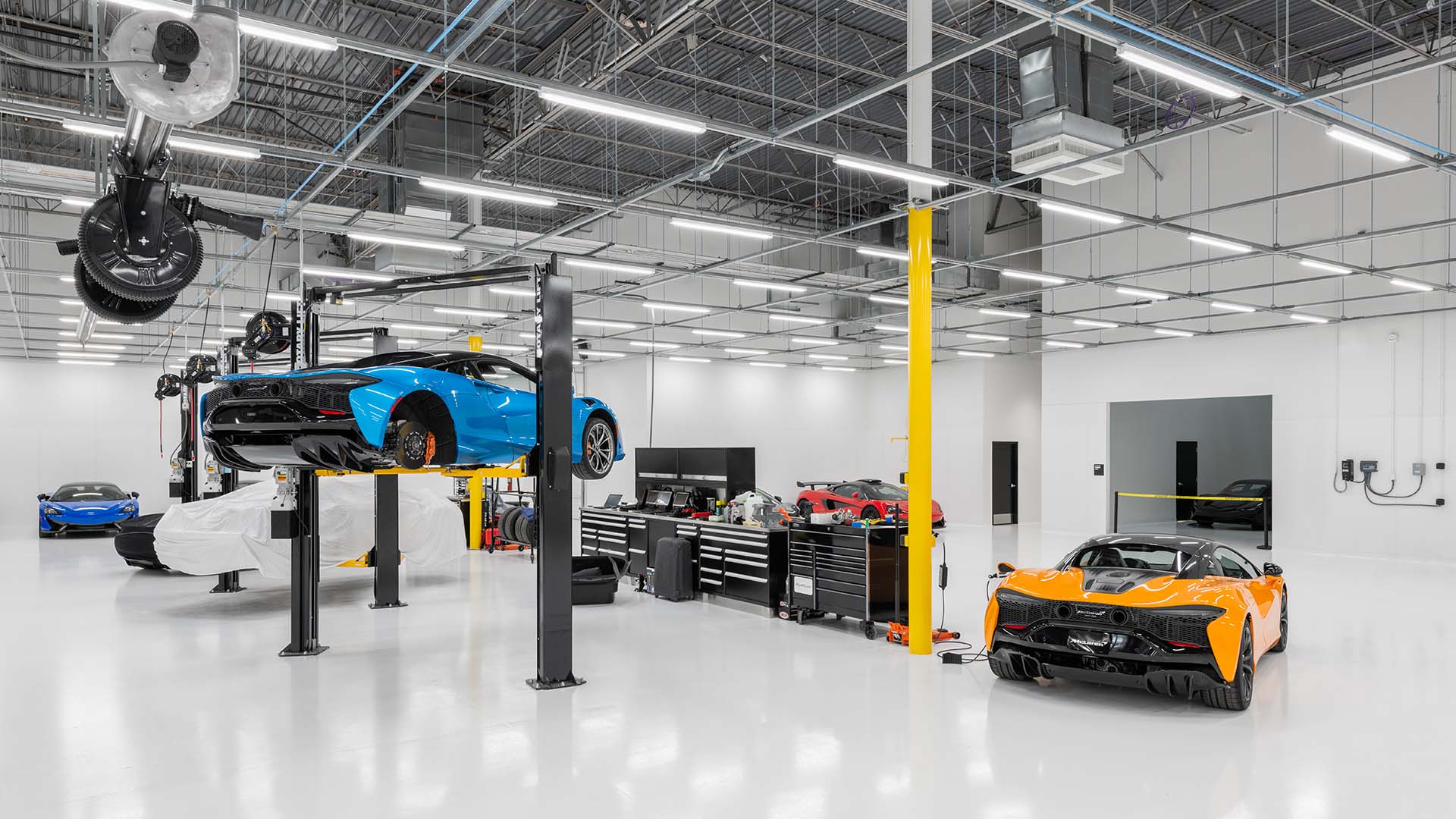 A clean, modern automotive workshop with luxury sports cars, including a blue car on a lift and an orange car parked on the white floor. Tool cabinets and equipment line the back wall.