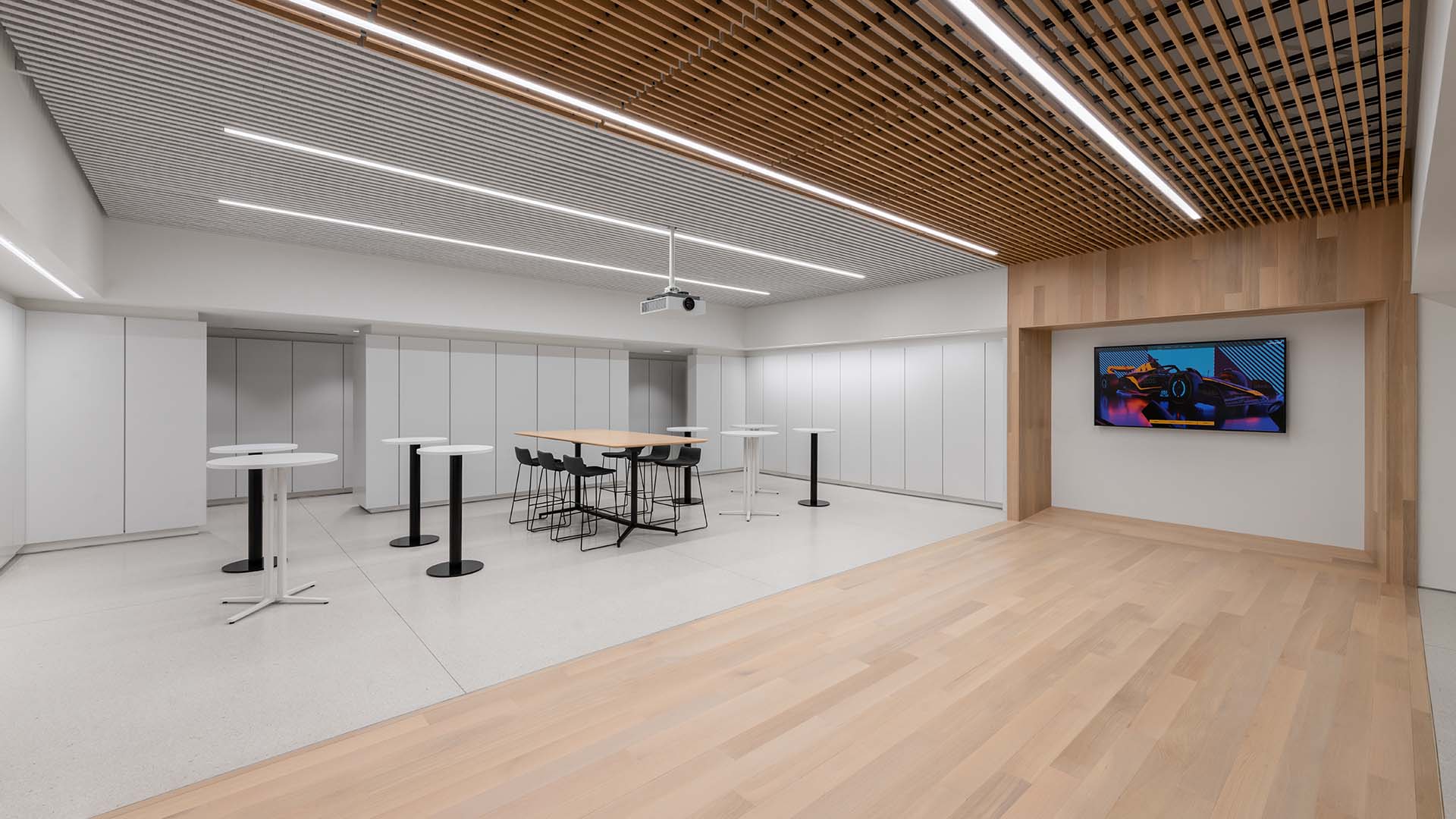 Modern conference room with white walls, high stools around a central table, small round tables, a ceiling-mounted projector, wood accents, and a wall-mounted display showing graphics.