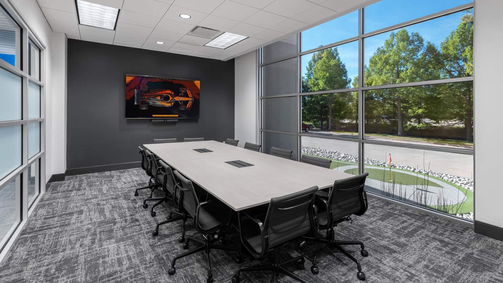 Modern conference room with a long table, ten black chairs, large windows with outdoor view, and a wall-mounted TV displaying a car image.