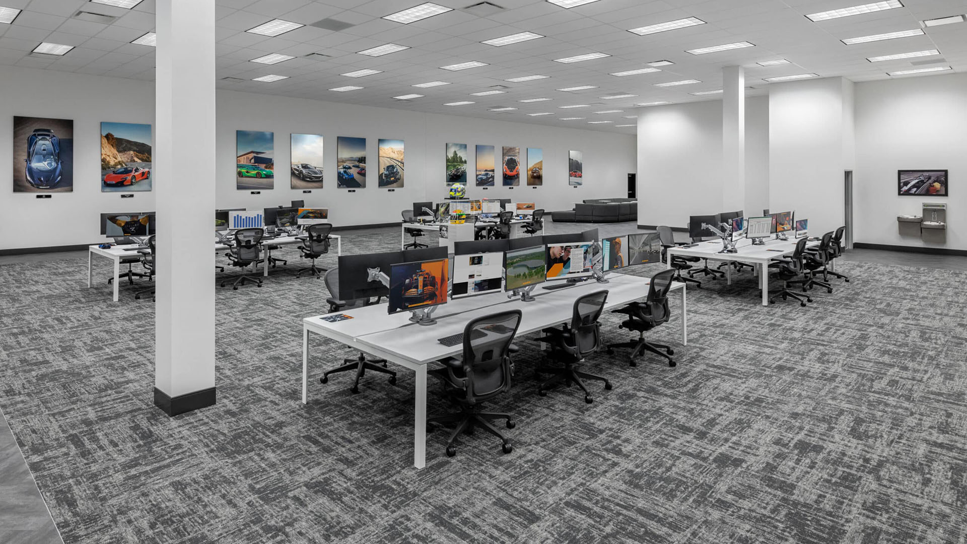 Modern open-plan office with rows of desks and computers, ergonomic chairs, carpeted floor, and automotive posters on the walls. The space appears organized and well-lit.