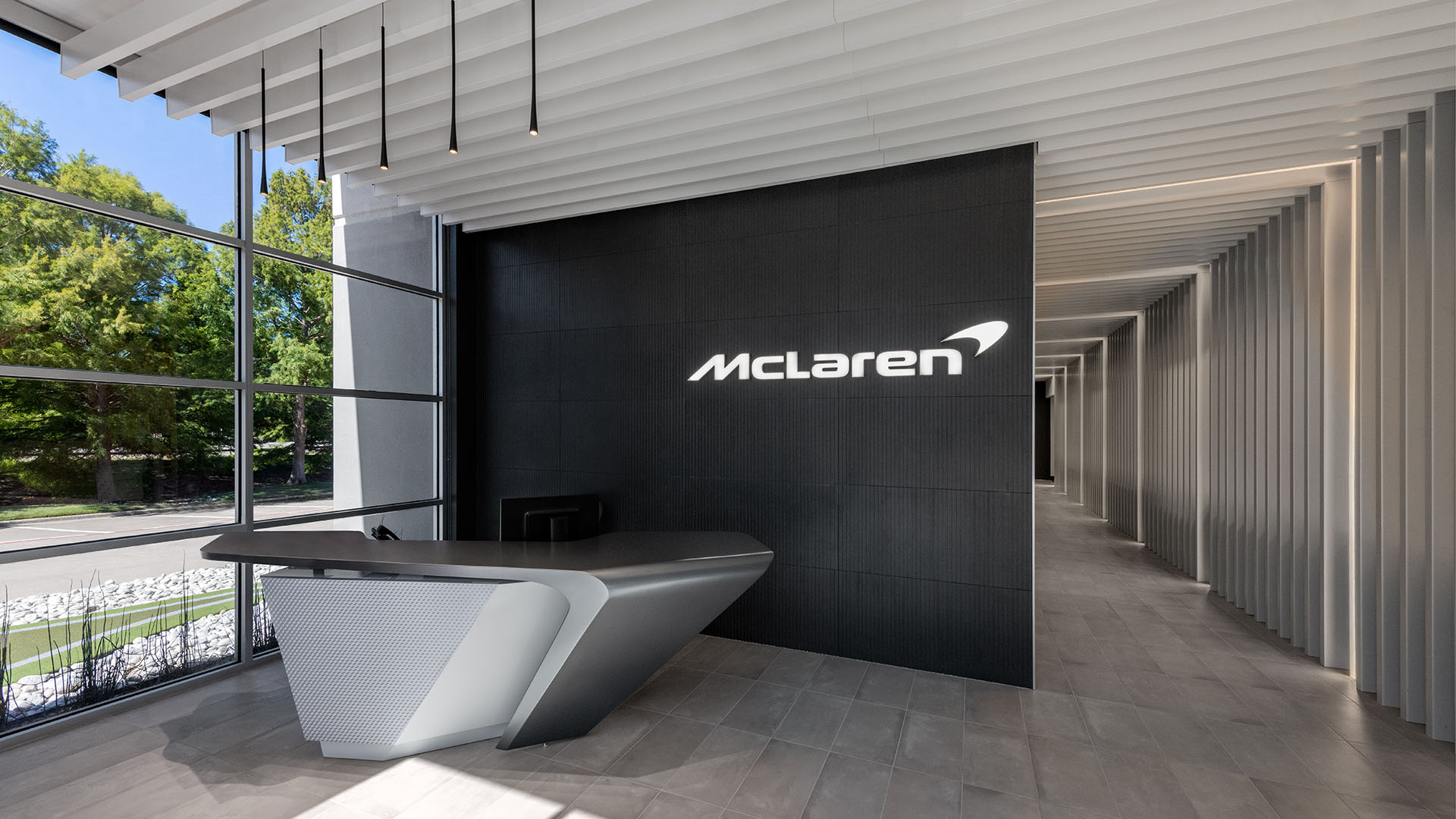 Modern reception area with a sleek desk, dark wall featuring a lit McLaren logo, and a corridor with white ceiling slats. Large windows reveal greenery outside.