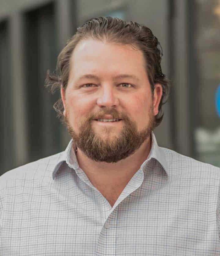 A man with a beard and mustache, wearing a light-colored checked shirt, stands outdoors in front of a blurred building background.