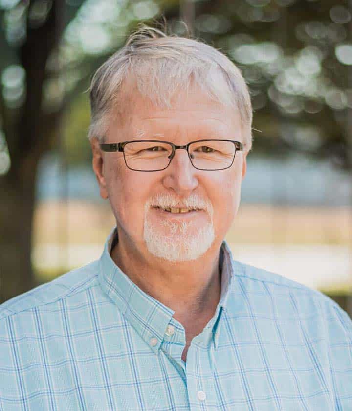 An older man with short gray hair, glasses, and a goatee smiles outdoors, wearing a light blue plaid shirt. Trees and sunlight are blurred in the background.