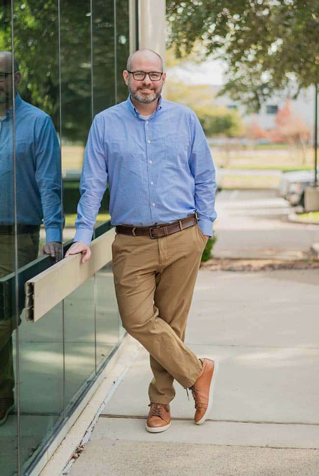 A man wearing a blue shirt and khaki pants stands outside, leaning against a glass wall with one leg crossed over the other.