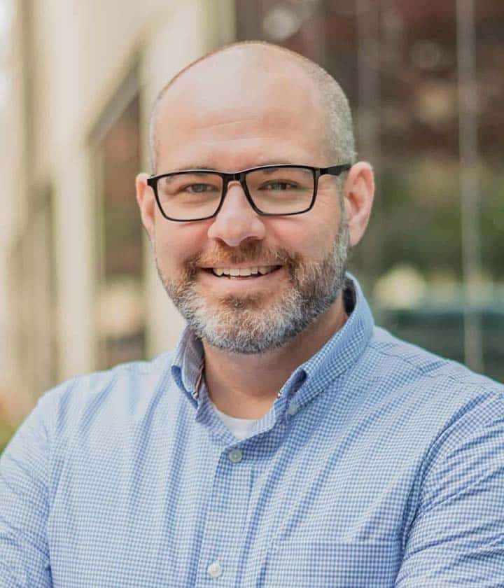 A bald man with a beard and glasses, wearing a blue checkered shirt, stands outdoors and smiles at the camera.