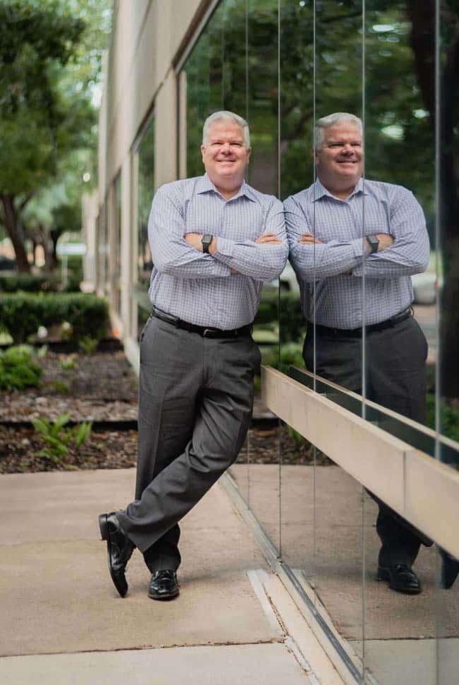 A man in business attire stands with arms crossed, leaning against a glass building, with his reflection visible in the window.