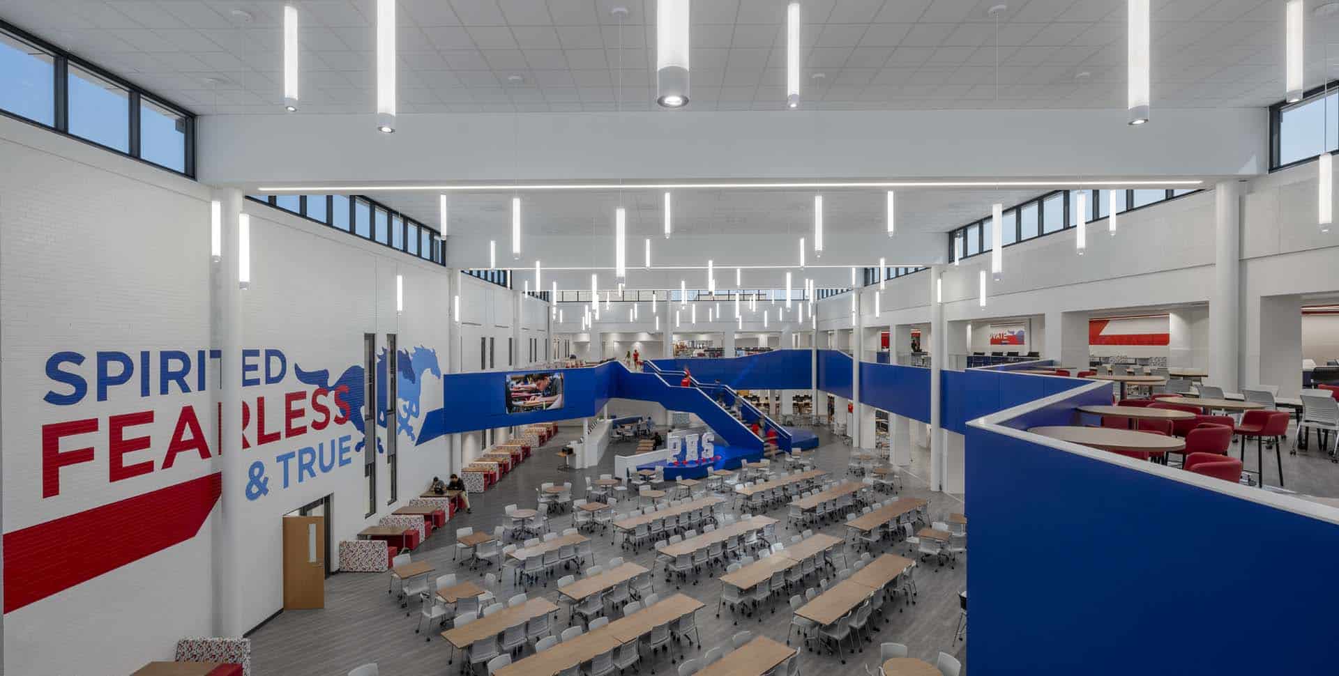Large, modern cafeteria with blue and white decor, rows of tables, a central staircase, and a mural reading "SPIRITED, FEARLESS & TRUE" on the wall.