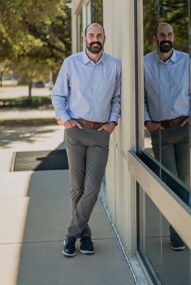 A man in a light blue shirt and gray pants stands with hands in pockets, leaning against a glass wall outside on a sunny day.