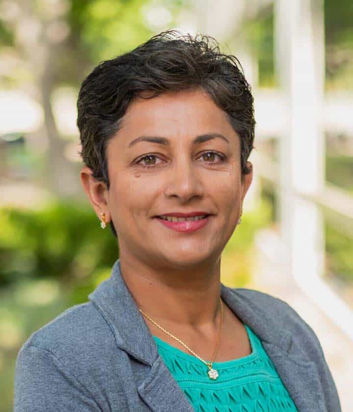 Woman with short dark hair wearing a gray blazer and teal top, smiling outdoors with greenery and blurred background.