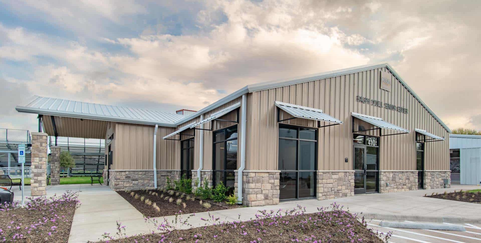 A modern, single-story building with metal siding, large windows, stone accents, and landscaped surroundings under a partly cloudy sky.