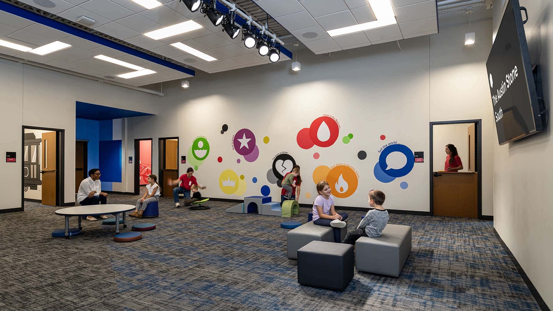 Children and adults are seated or playing in a modern, colorful classroom with geometric wall designs and bright lighting.