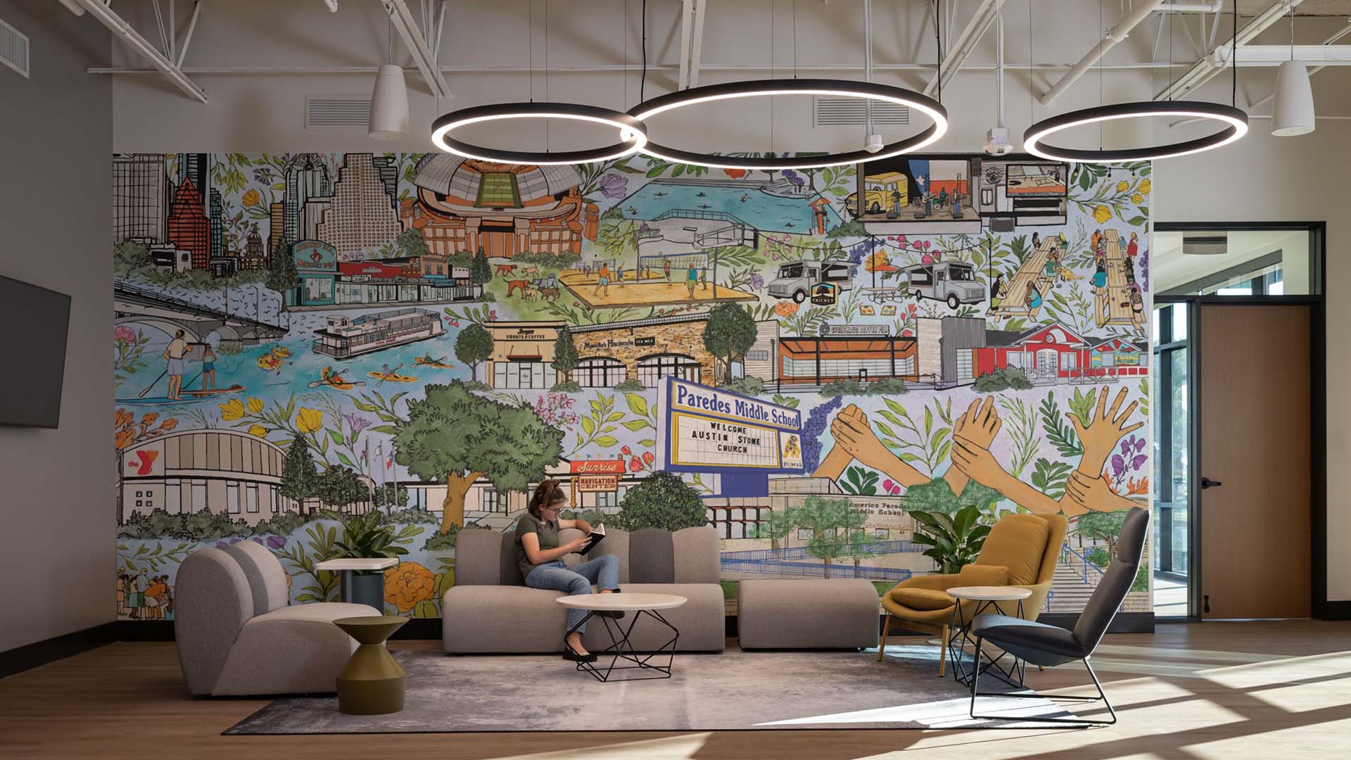A woman sits on a sofa in a modern lounge area with a large, colorful mural depicting various scenes and landmarks on the wall behind her.