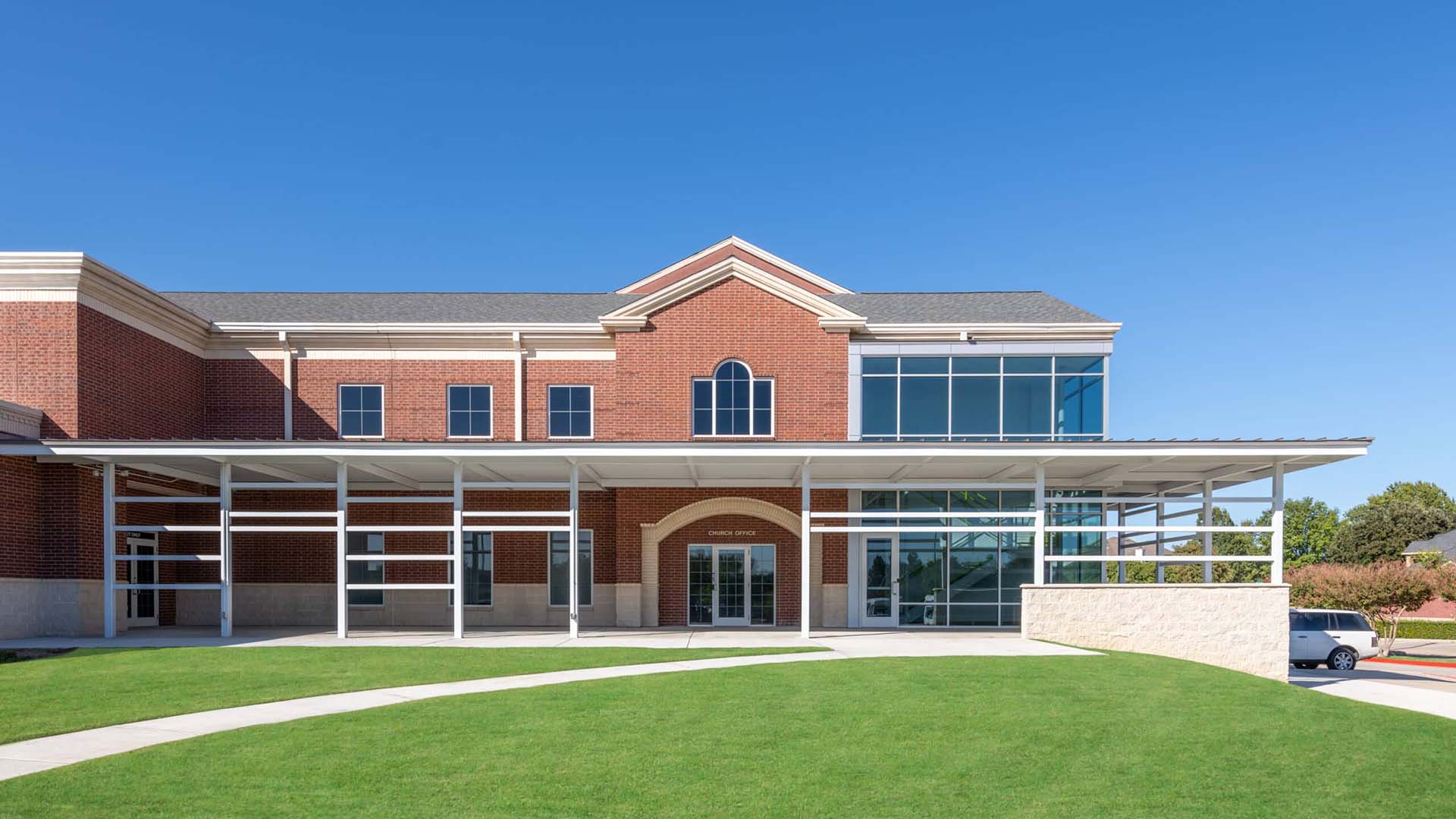 A modern red brick building with large glass windows, a covered entrance, and a well-kept lawn under a clear blue sky.