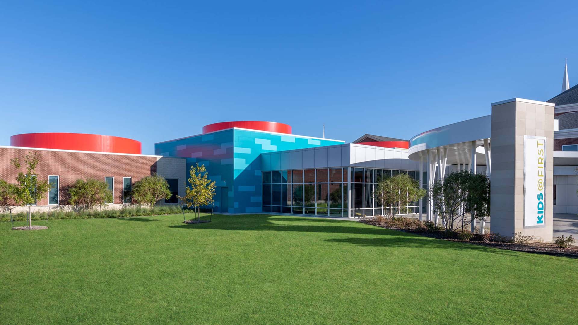 Modern building with blue and red accents, large windows, a "KIDS FIRST" sign, and a green lawn in front under a clear blue sky.