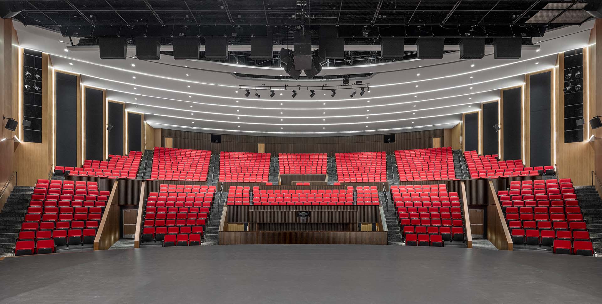 A modern theater auditorium with red seats arranged in tiers, wood paneling on the walls, and overhead stage lighting.