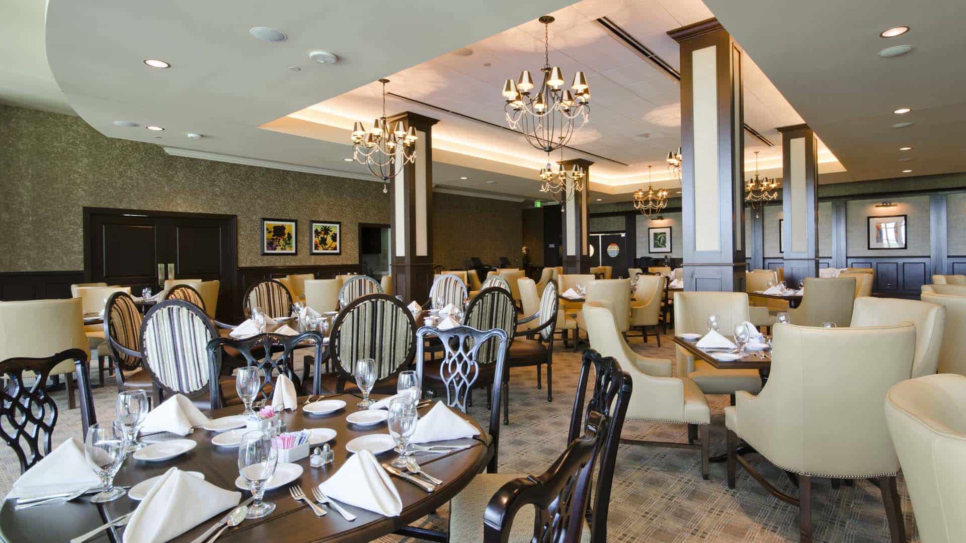 Elegant restaurant interior with round tables set with white napkins and glassware, upholstered chairs, chandeliers, and framed artwork on the walls.