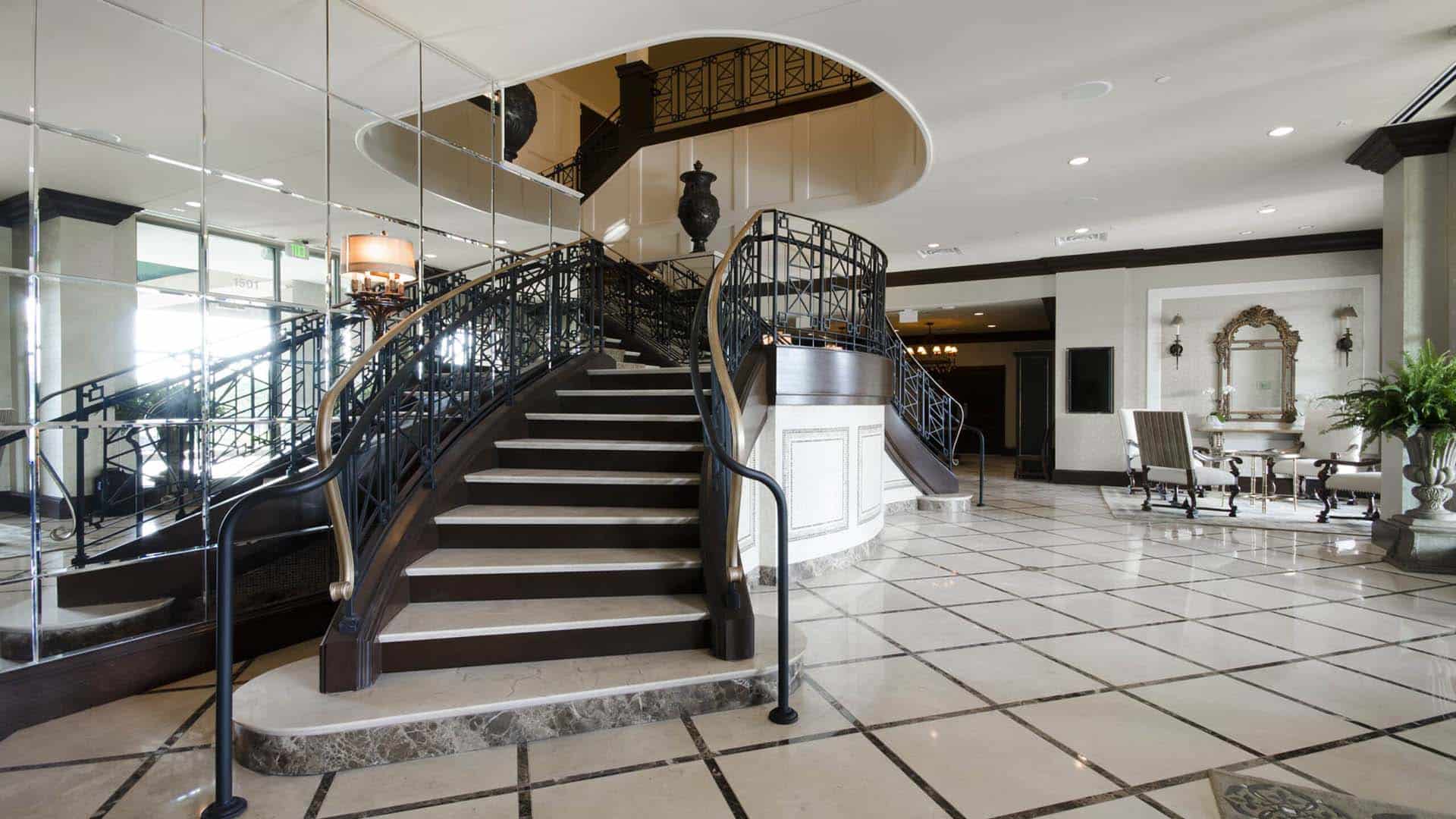 Grand staircase with black railing and marble steps in a spacious, elegant lobby with tiled floor, mirrored walls, and seating area in the background.