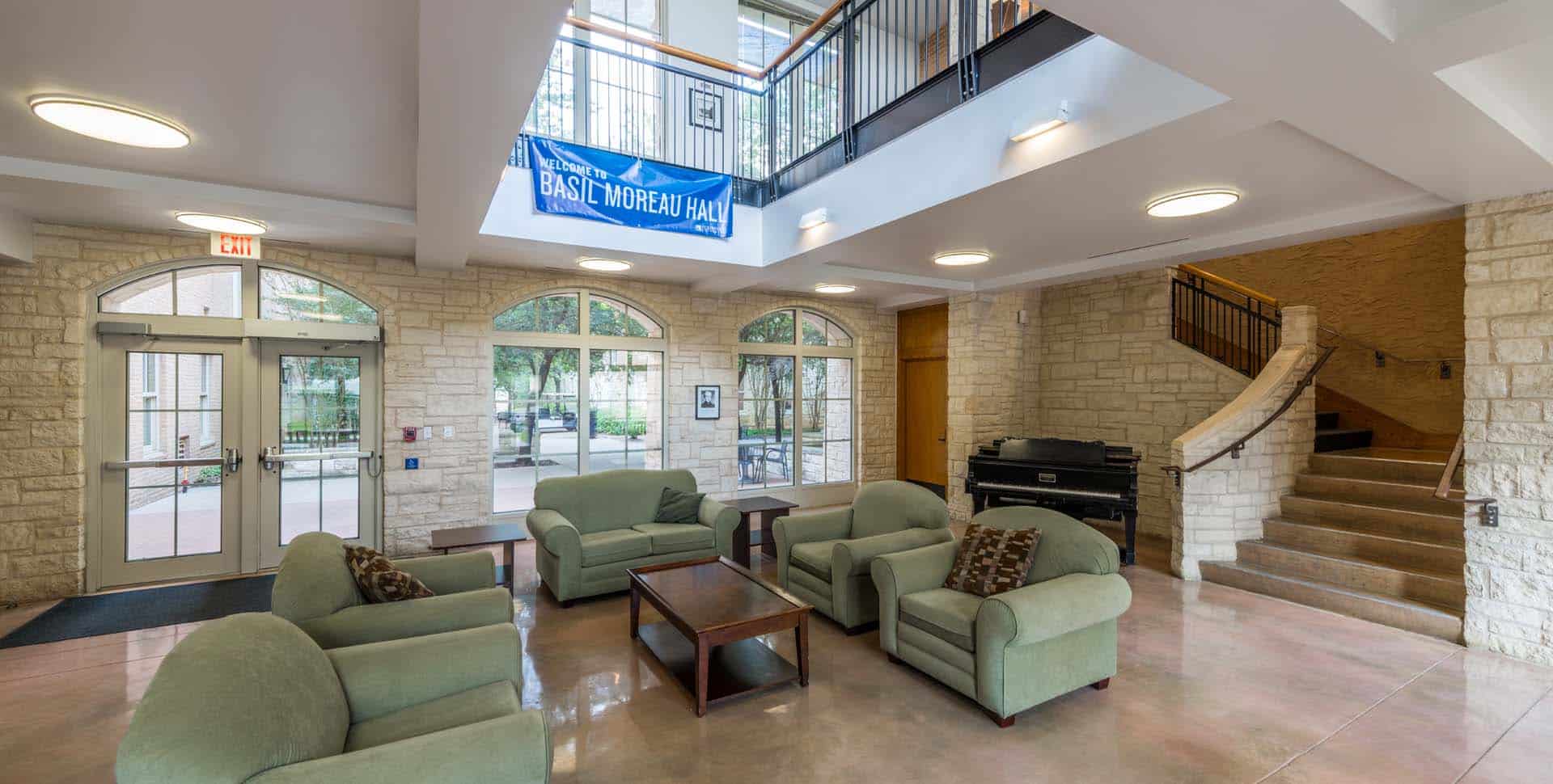 A lounge area with green armchairs, a coffee table, stone walls, a piano, a staircase, and a banner reading "Basil Moreau Hall" hanging from the upper level.