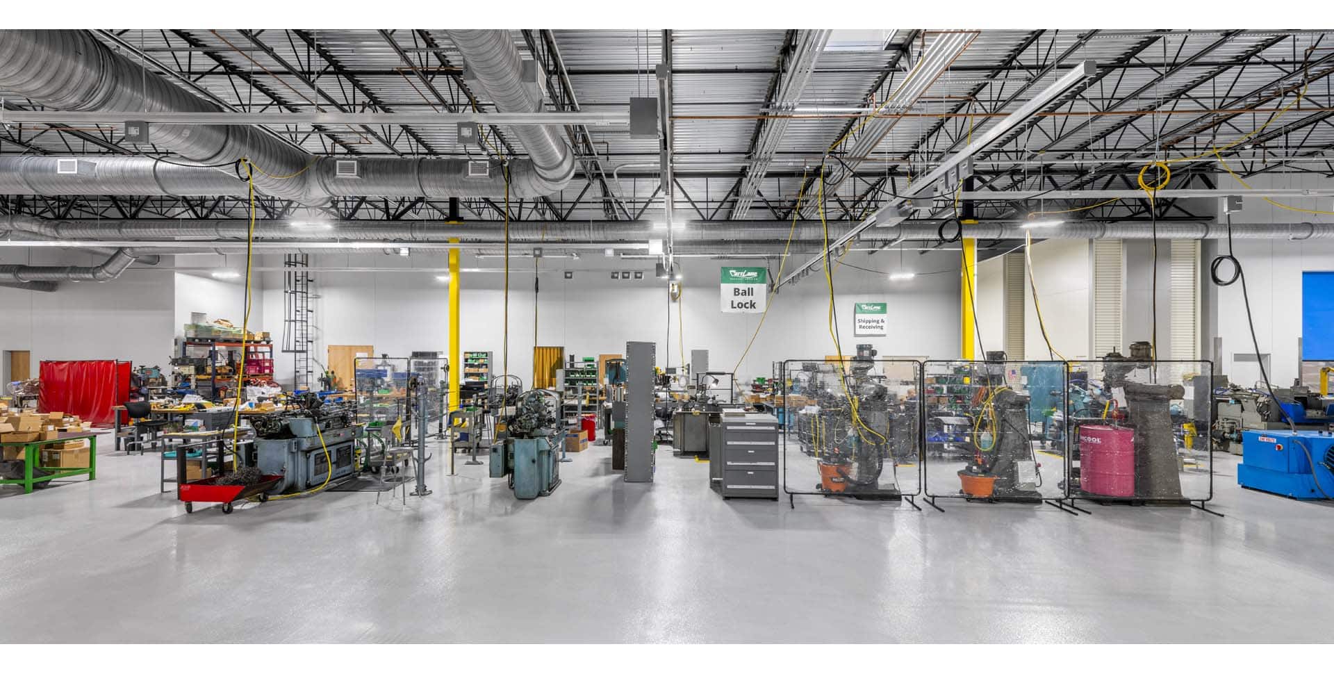 A spacious industrial workshop with various machines, workstations, tools, and safety barriers under bright overhead lighting and exposed ceiling ducts.
