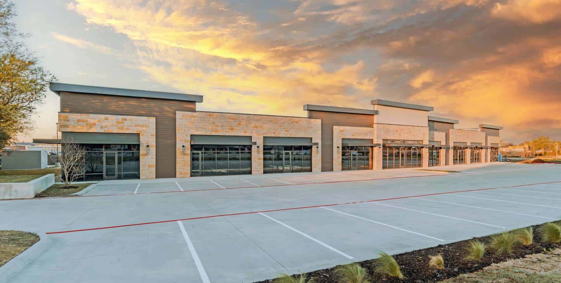 A row of empty, modern retail storefronts with large windows and stone accents, set against a dramatic sunset sky in a vacant parking lot.
