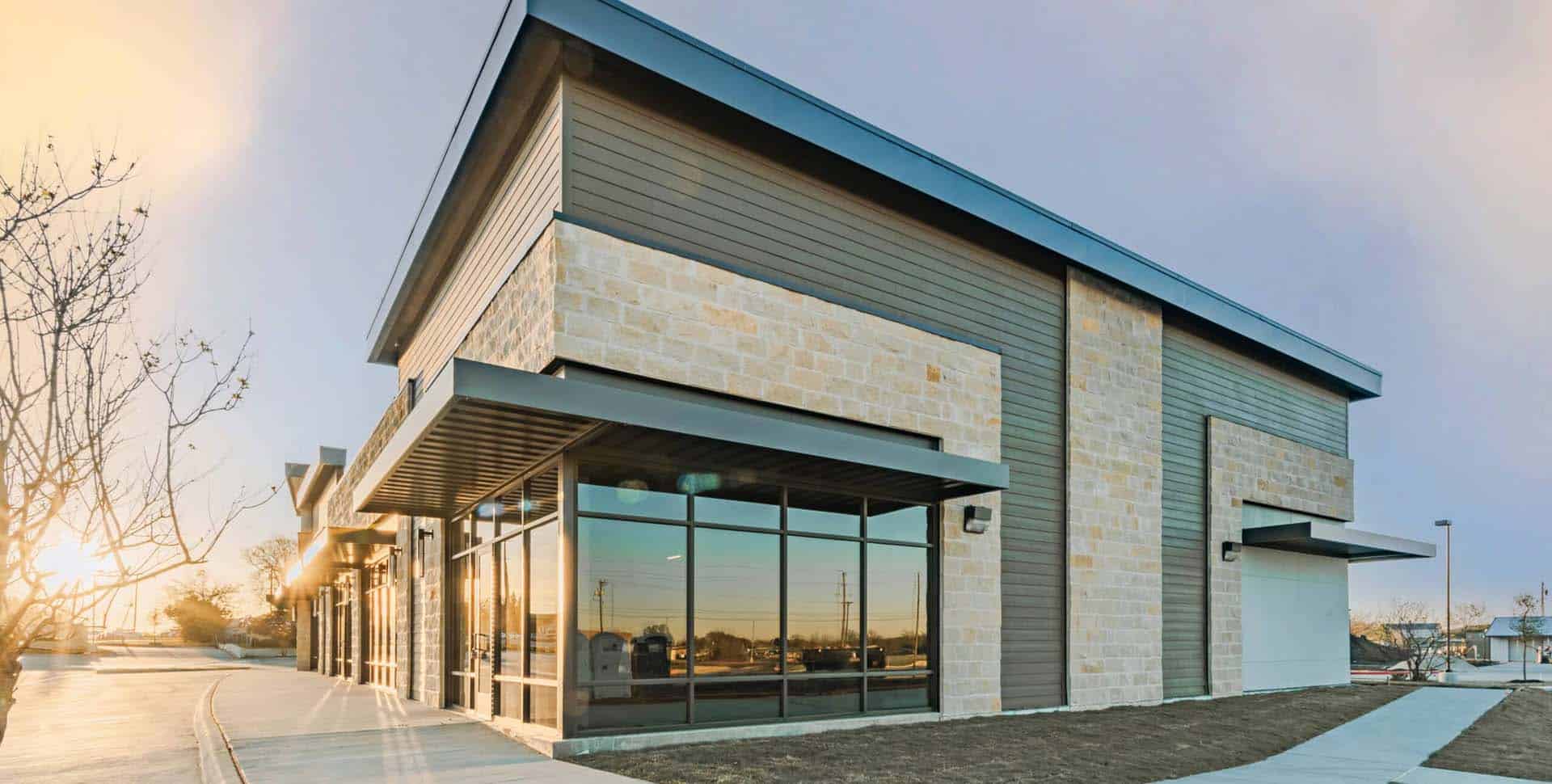 Modern commercial building with large glass windows, stone and metal facade, seen at sunset with a clear sky and sidewalk in the foreground.