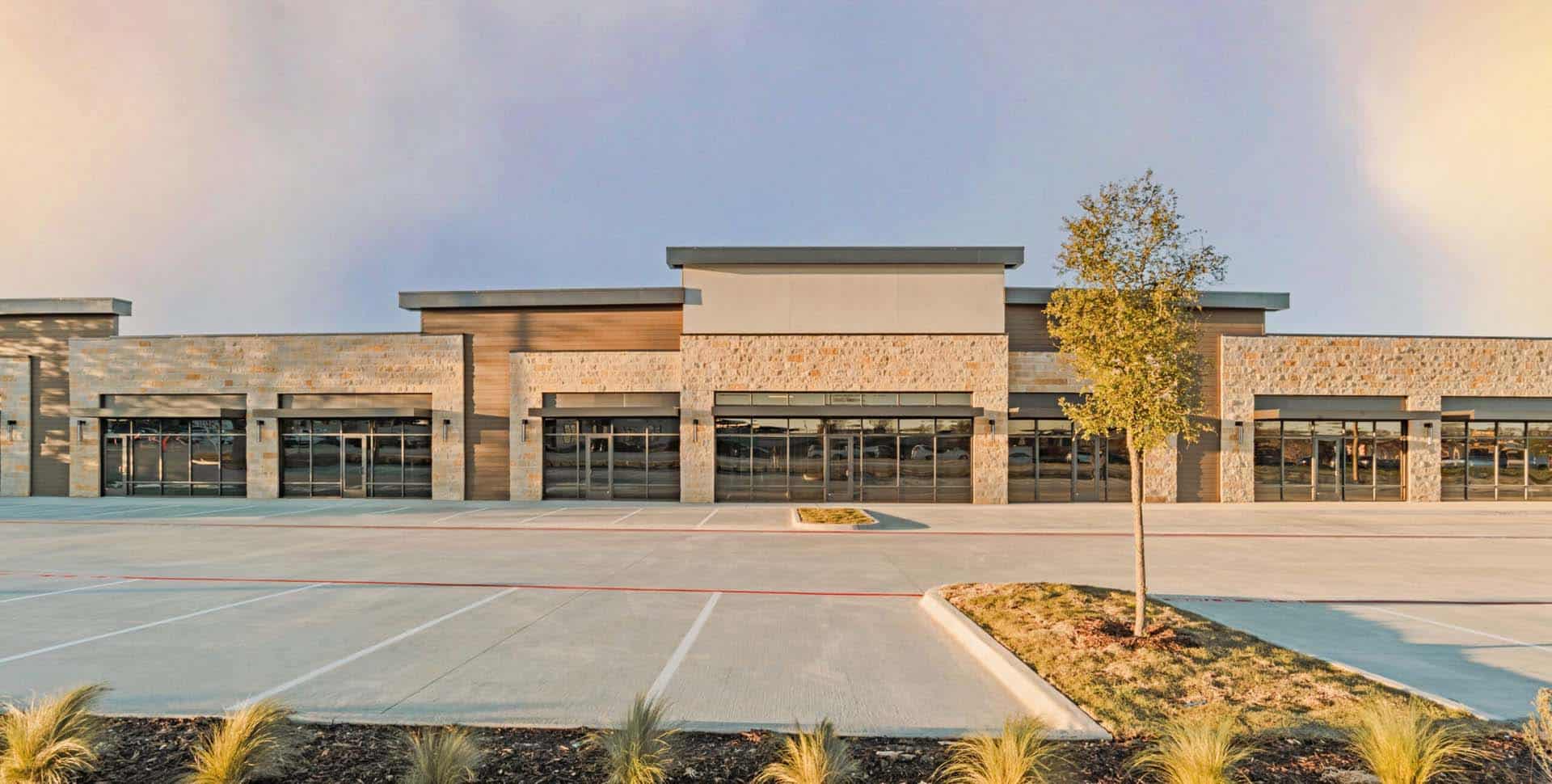 A modern, empty strip mall with stone facade, large glass windows, and an empty parking lot in front, featuring a small tree and landscaped area.
