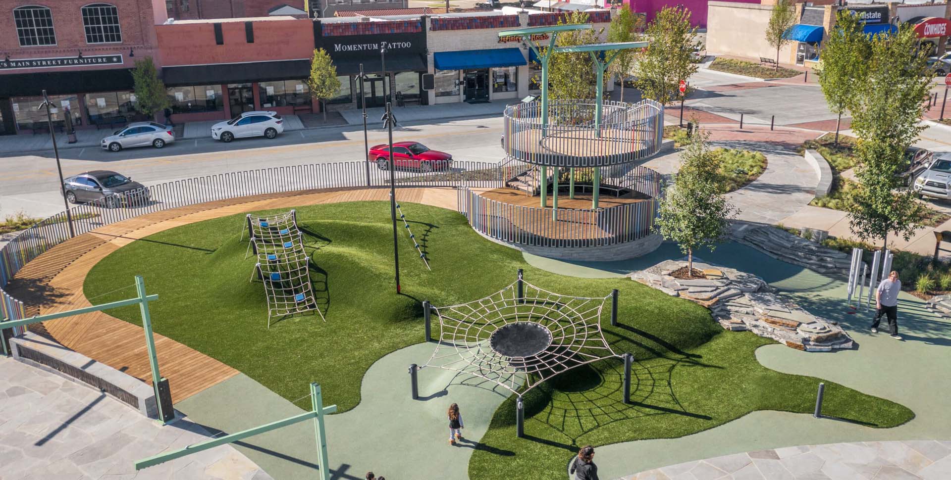 A modern playground with climbing structures, rope nets, and a circular elevated platform, bordered by a street with parked cars and shops.