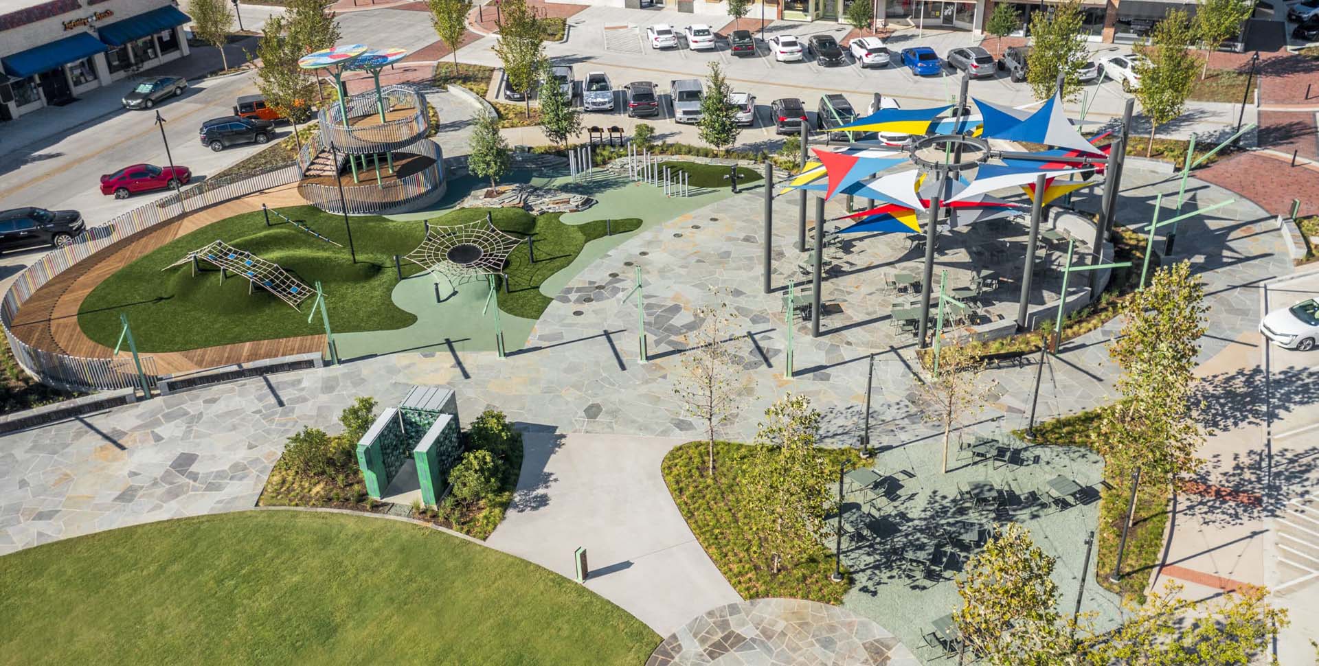 Aerial view of a modern outdoor playground with climbing structures, artificial turf, shaded seating areas, nearby shops, and parked cars.