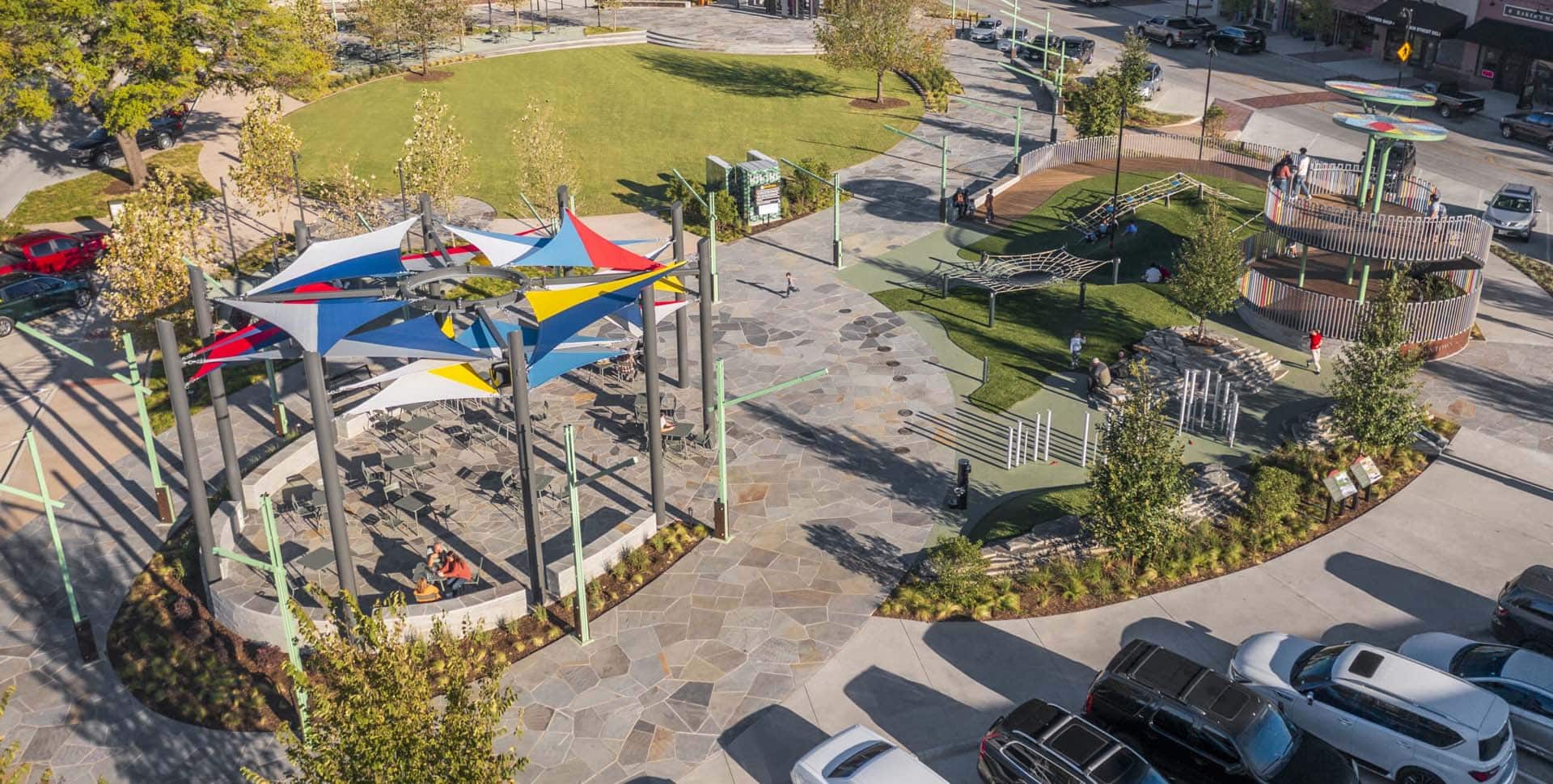 Aerial view of a public park with a shaded seating area, playground equipment, green lawn, and parked cars nearby.