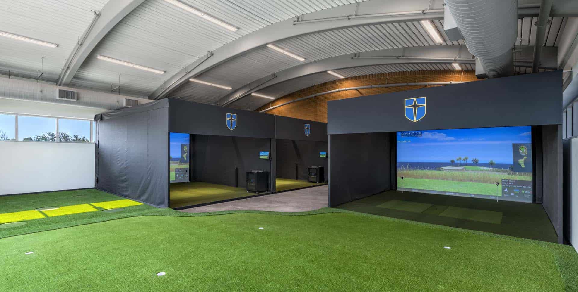 Indoor golf facility featuring two golf simulators with large screens and a putting green in the foreground, under an arched ceiling.