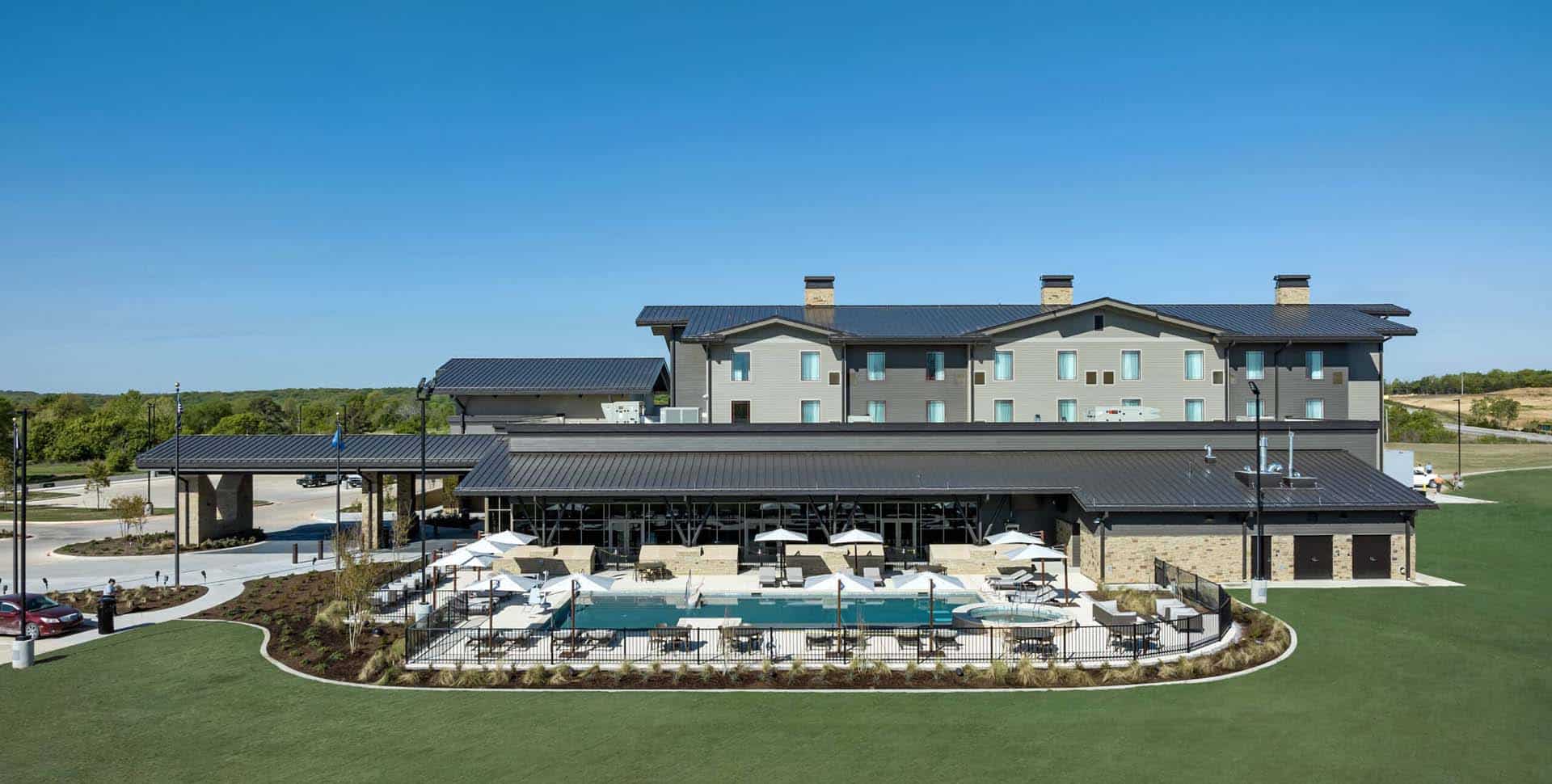 A modern three-story building with a pool, lounge chairs, and umbrellas in front, surrounded by landscaped grounds under a clear blue sky.