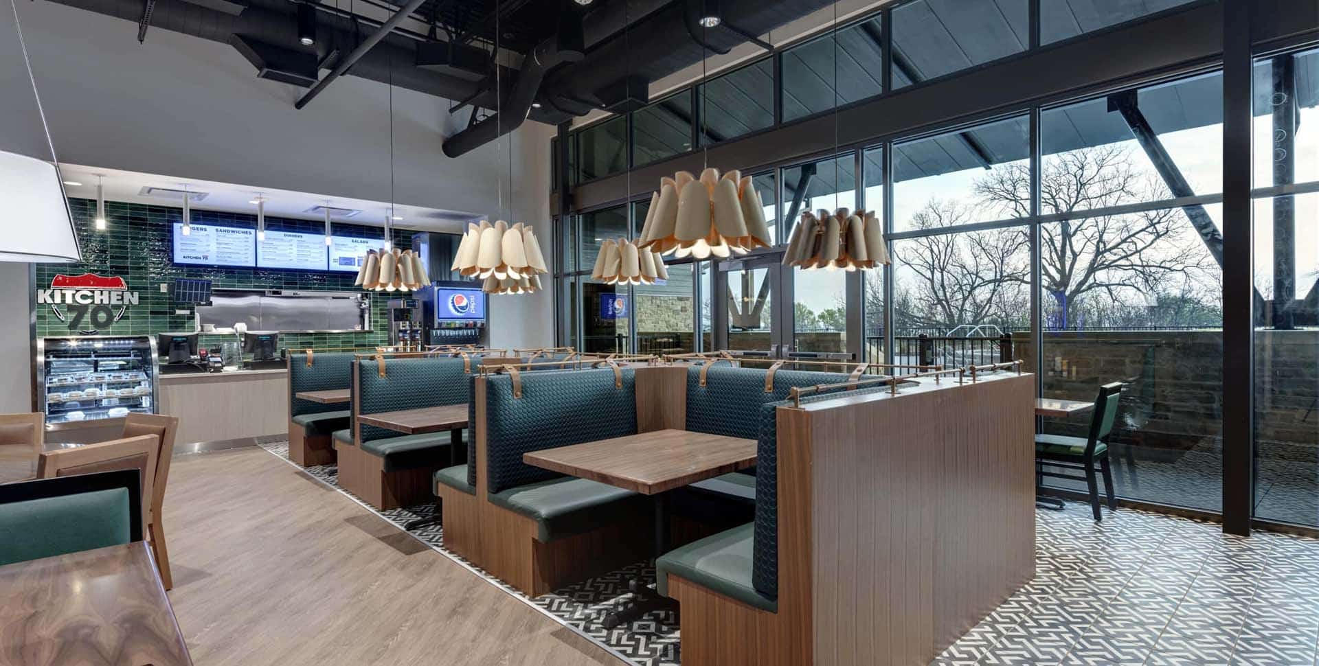 Modern fast-casual restaurant interior with green booth seating, wood tables, pendant lights, menu screens, and large windows showing trees outside.