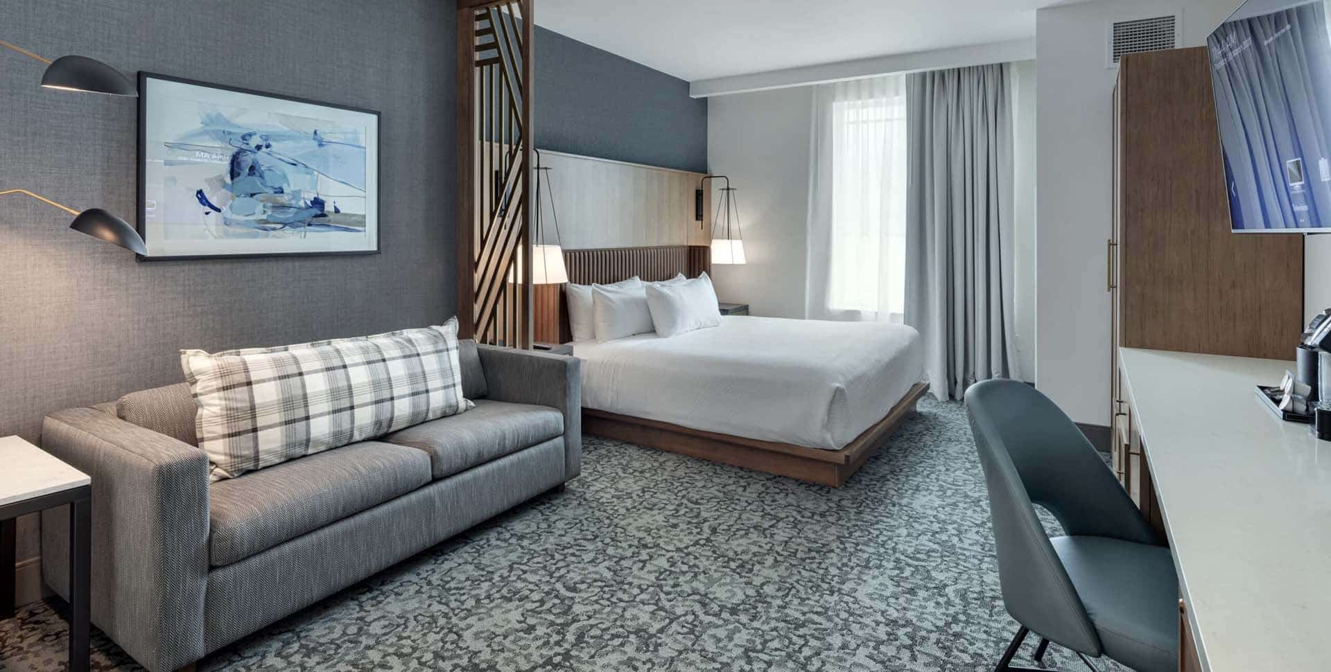 Modern hotel room with a neatly made bed, gray sofa, desk with chair, large window with sheer curtains, and abstract artwork on the wall.